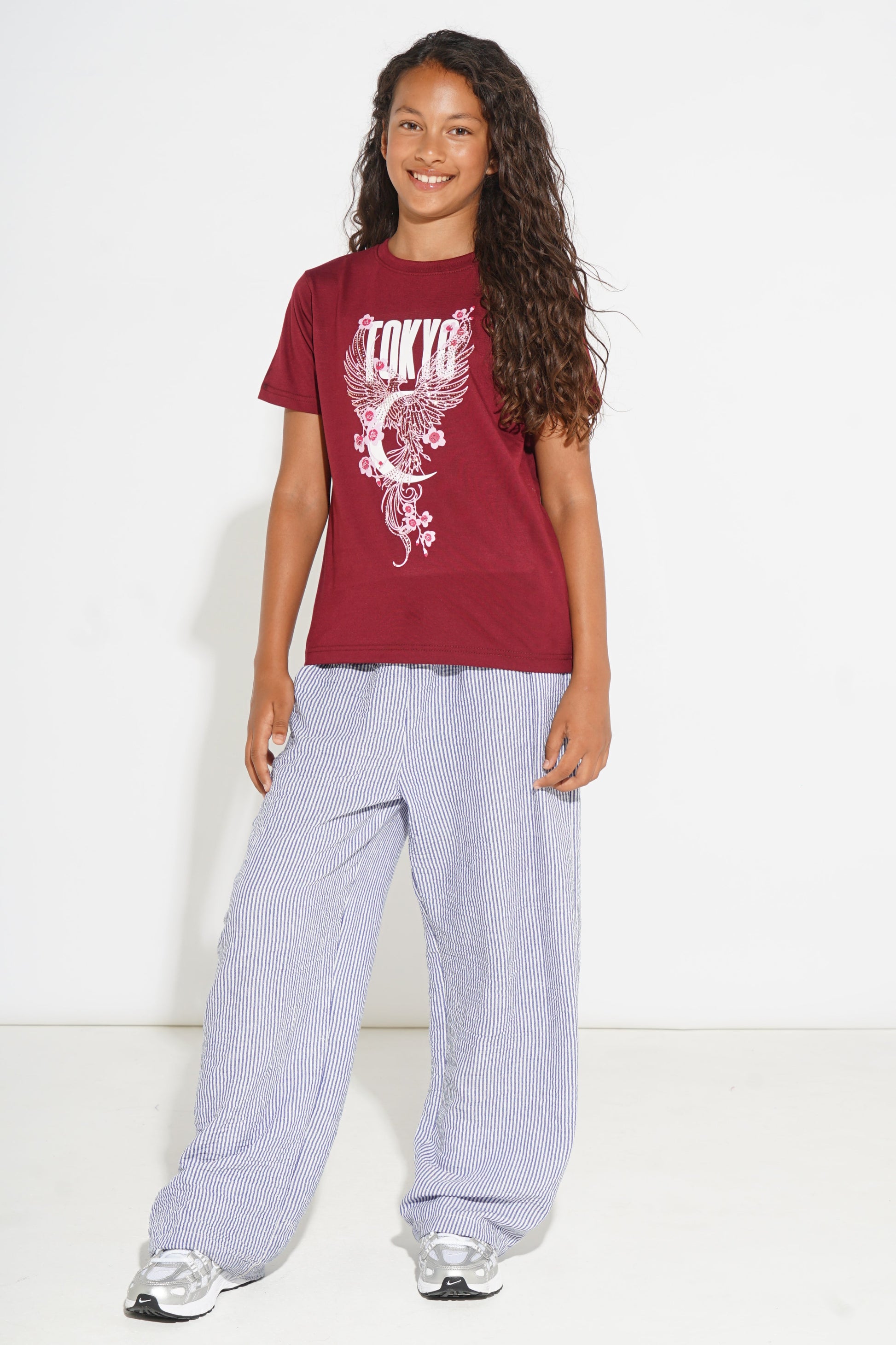 A young girl with long curly brown hair stands against a white studio background wearing a maroon short-sleeve graphic T-shirt with a white 'Tokyo' dragon print, loose light purple wide-leg pants, and silver sneakers.