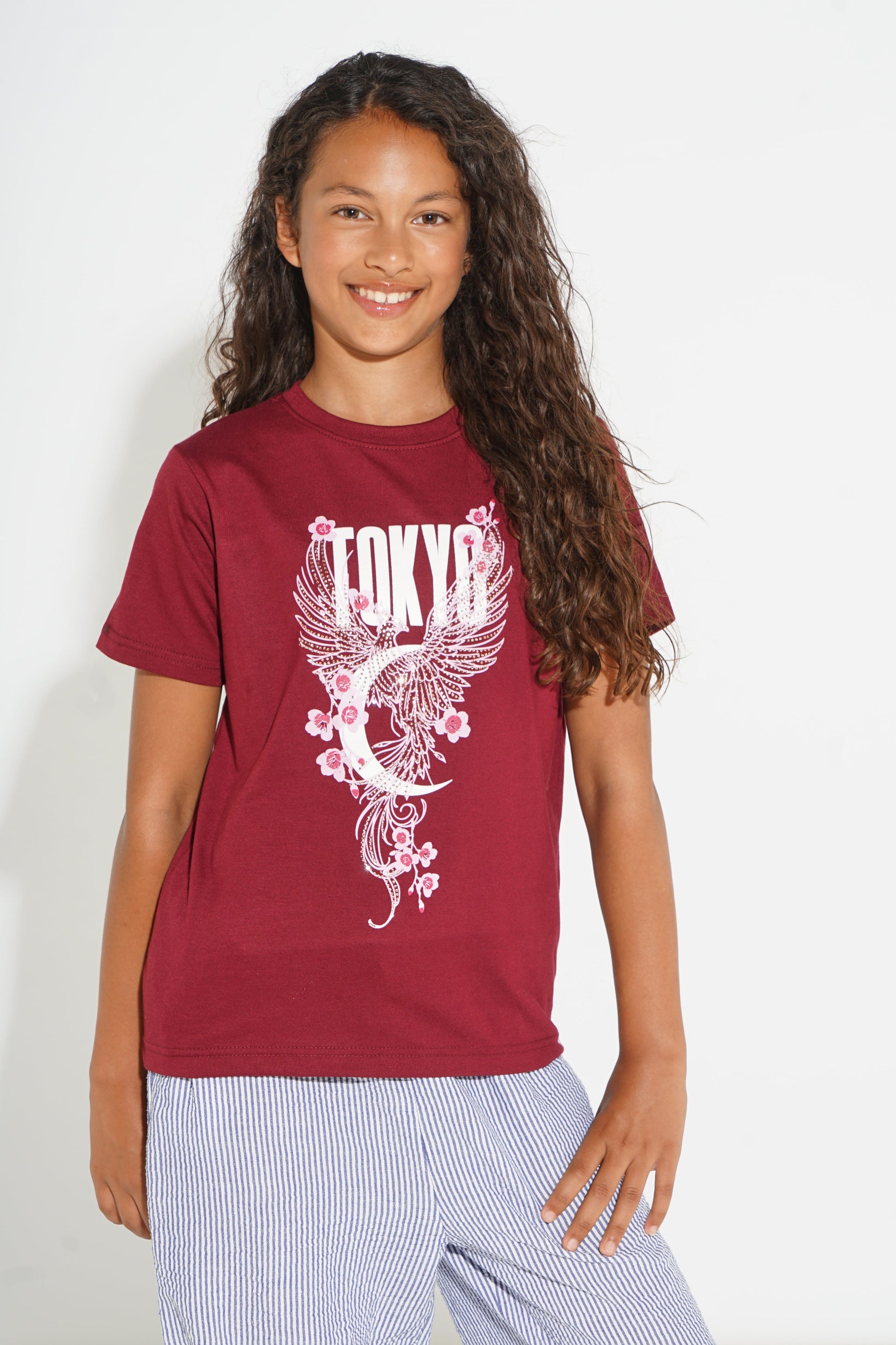 A young girl with long, curly brown hair smiling, wearing a maroon short-sleeve graphic t-shirt with a white 'Tokyo' crescent moon and cherry blossom design, paired with light blue and white striped pants, against a plain white background.