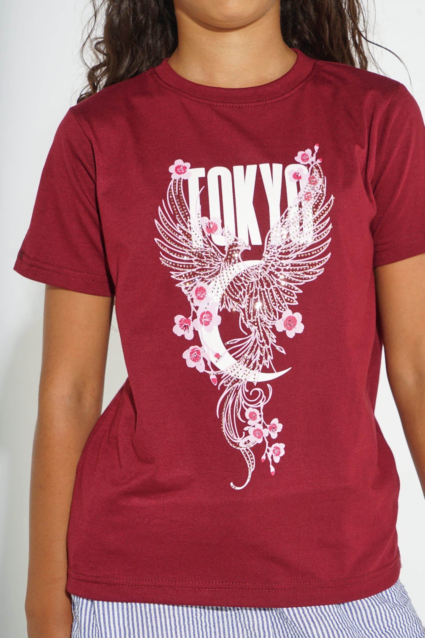 A person wearing a short-sleeve crewneck t-shirt in maroon red, featuring a white graphic print of a phoenix, crescent moon, cherry blossoms, and the word 'TOKYO'.