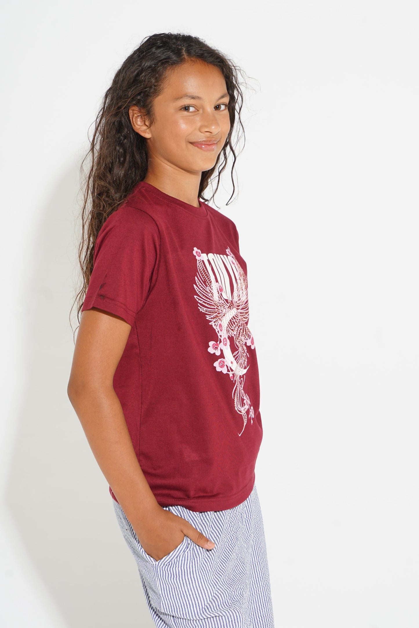 A young girl with long dark curly hair is shown from the waist up, wearing a maroon short-sleeve t-shirt with a white graphic print of a bird and stripes, paired with light blue and white striped pants, against a plain white background.