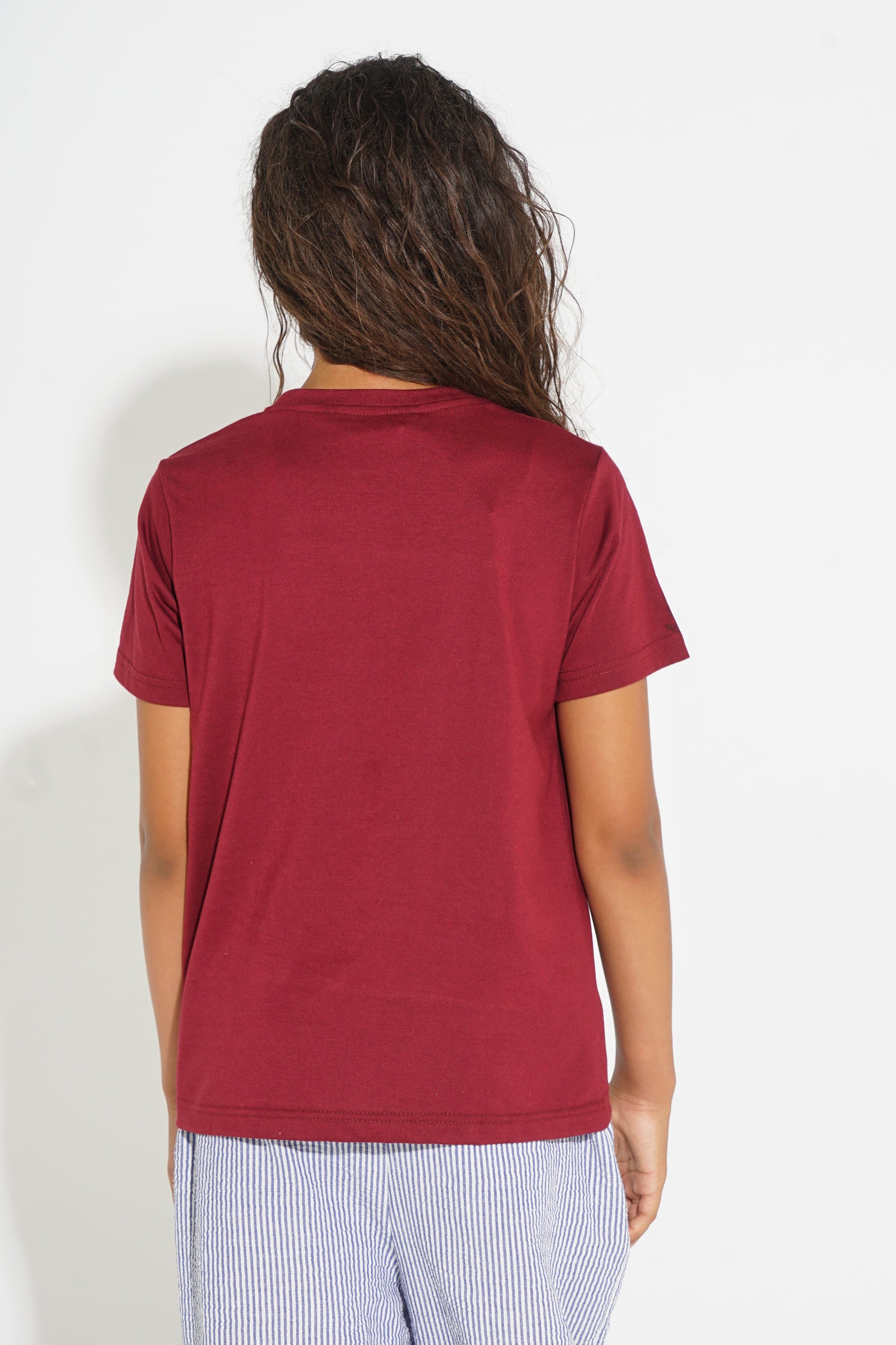 A person shown from the back wearing a short-sleeve crew-neck t-shirt in a deep burgundy red, paired with light blue and white striped pants, against a plain white background.