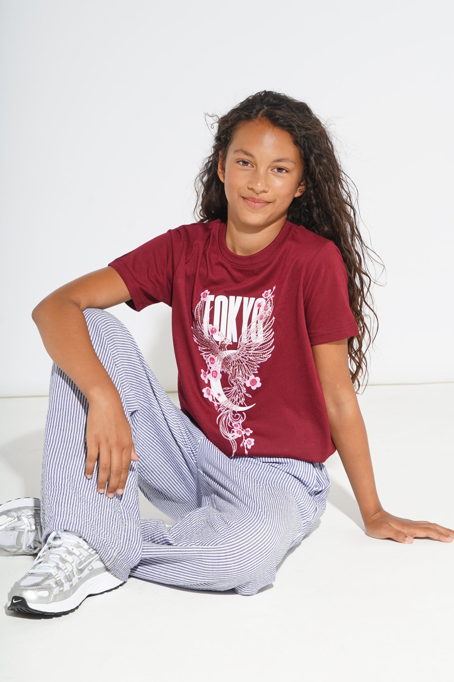 A young girl with long curly hair sits on a white floor against a white wall, wearing a maroon graphic t-shirt with a white 'Tokyo' dragon design and light blue and white striped pants.