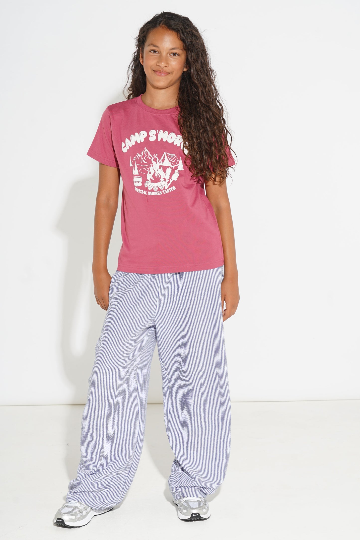 A girl modeling a pink short-sleeve t-shirt with a white 'CAMP SMOKE' graphic, paired with light blue wide-leg pants and white sneakers, standing against a white studio background.