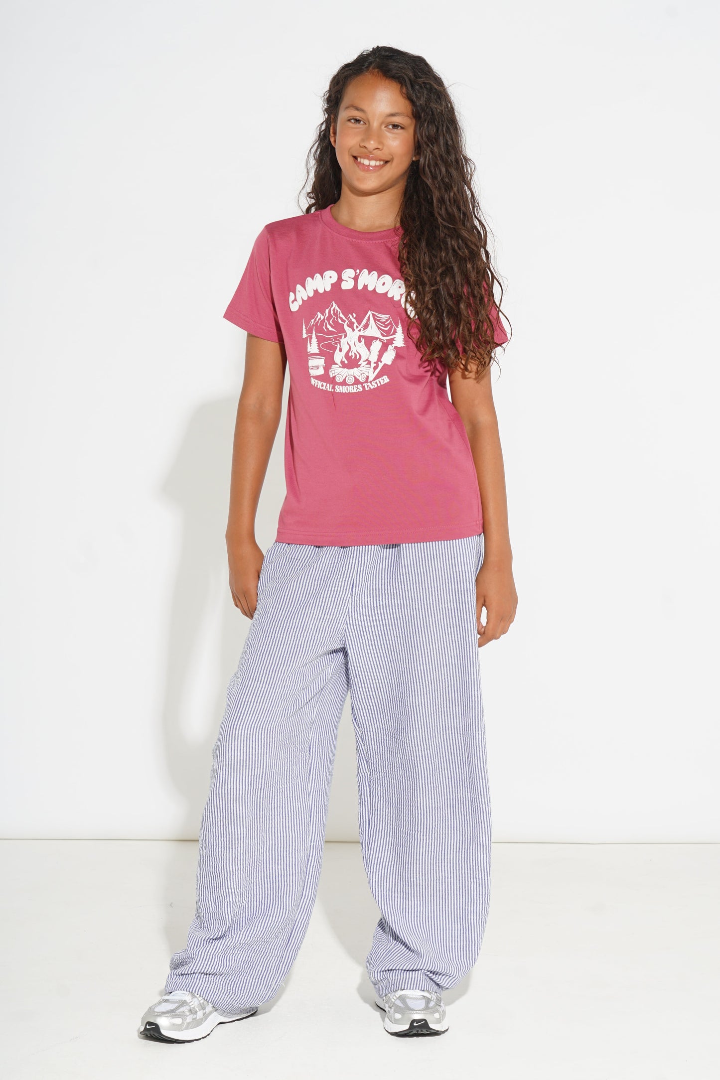 A young girl with long curly brown hair stands against a white studio background wearing a dusty rose pink graphic t-shirt with 'CAMP SMOKE' printed in white, paired with wide-leg light purple trousers and white sneakers.