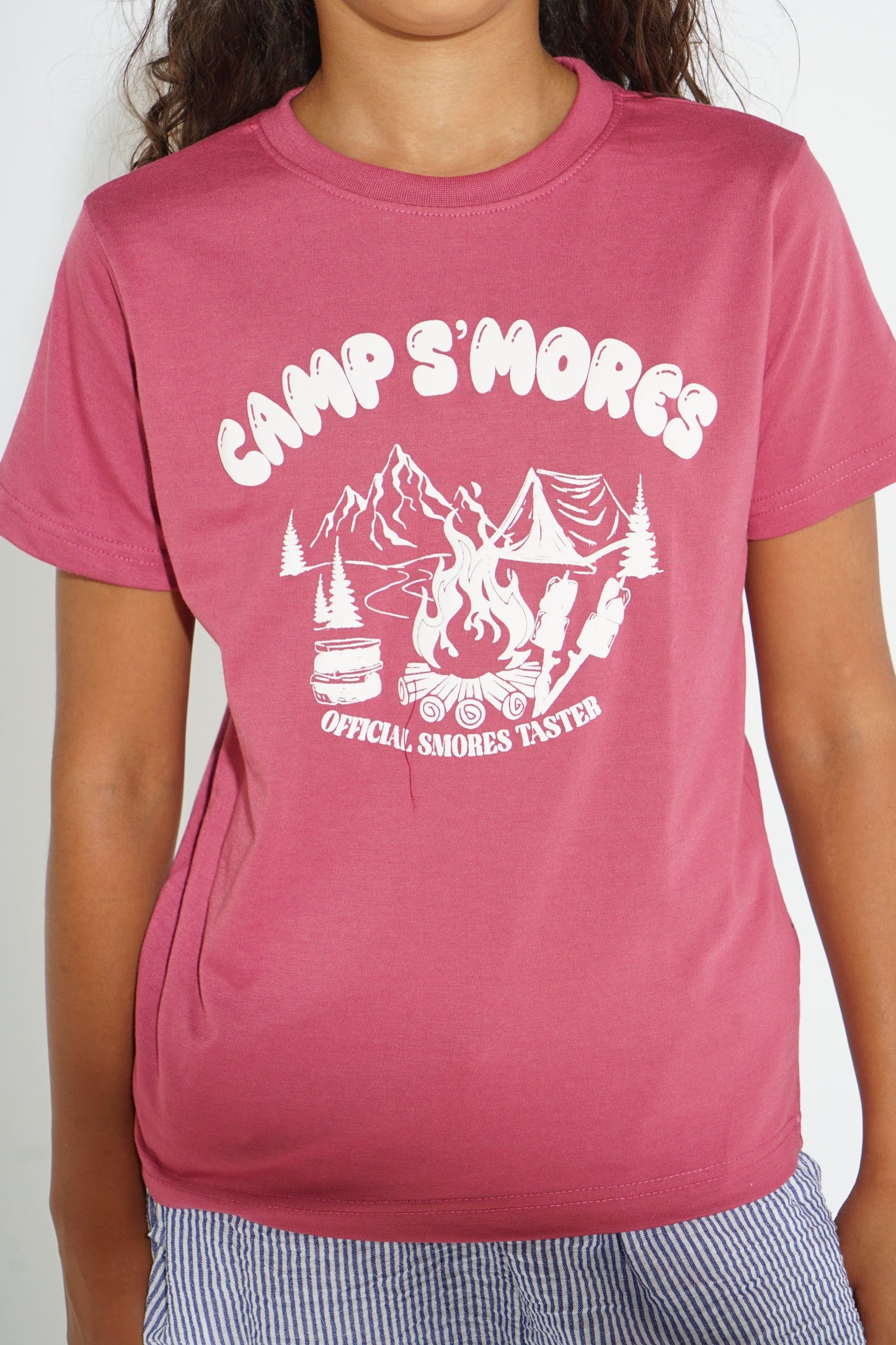 A person wearing a pink short-sleeve crewneck t-shirt with a white graphic print that reads 'CAMP S'MORES' and 'OFFICIAL SMOORES TASTER' with a campfire and mountains design.