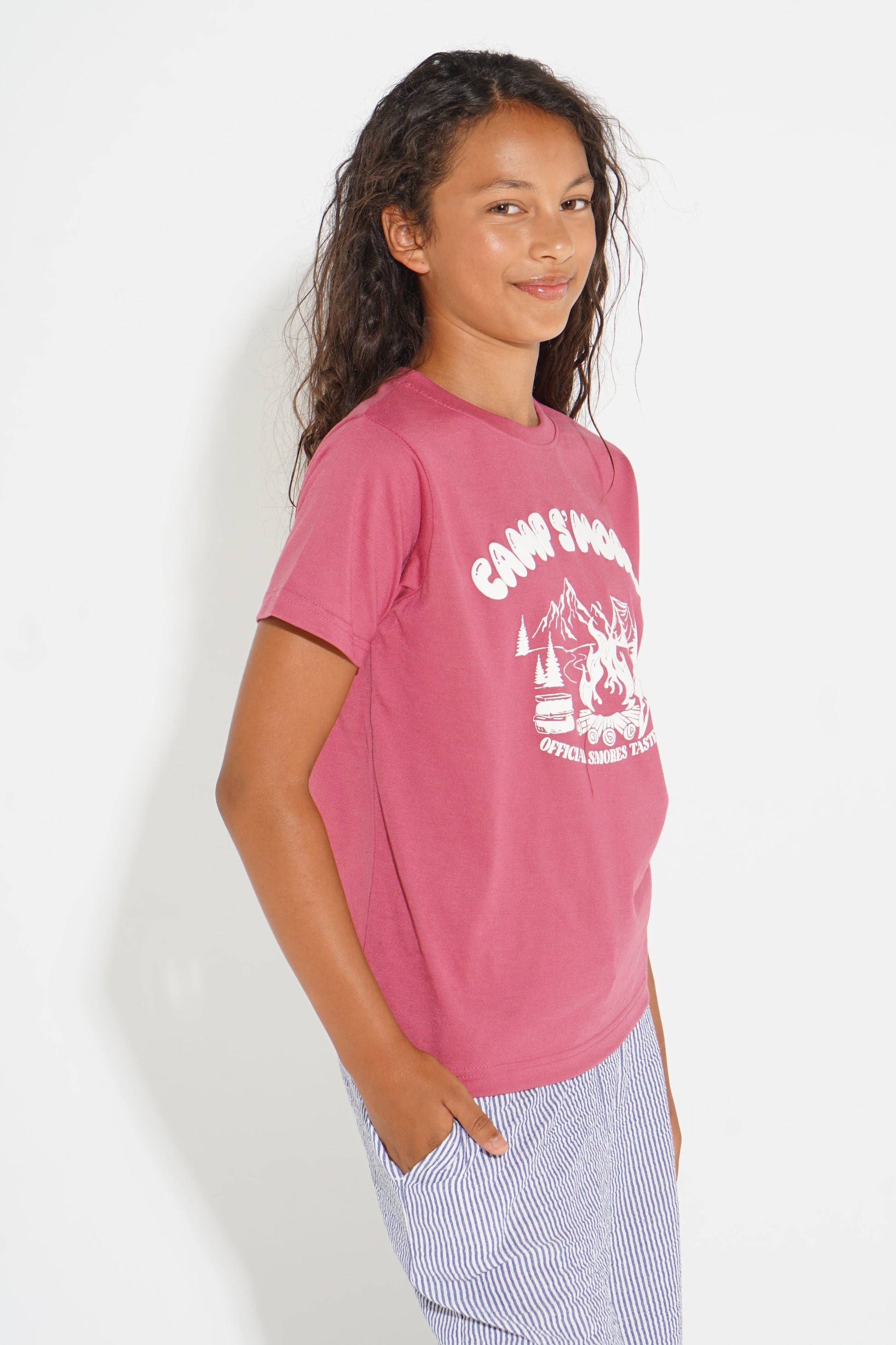 A young girl with long dark curly hair is shown from the side, wearing a pink short-sleeve t-shirt with a white graphic print that reads 'CAMP FINDER' and 'OFFICE OF MYTHS', paired with light blue and white striped pants, against a plain white background.