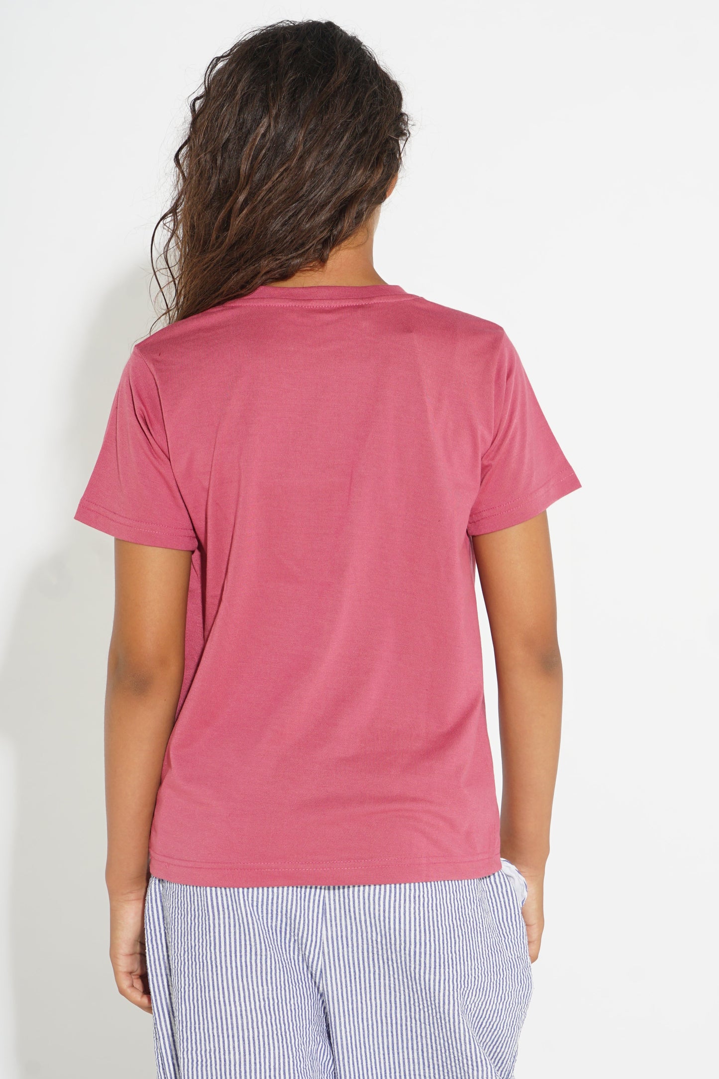 A woman viewed from the back wearing a short-sleeve crewneck t-shirt in a dusty rose pink color, paired with light blue and white striped pants, against a plain white background.
