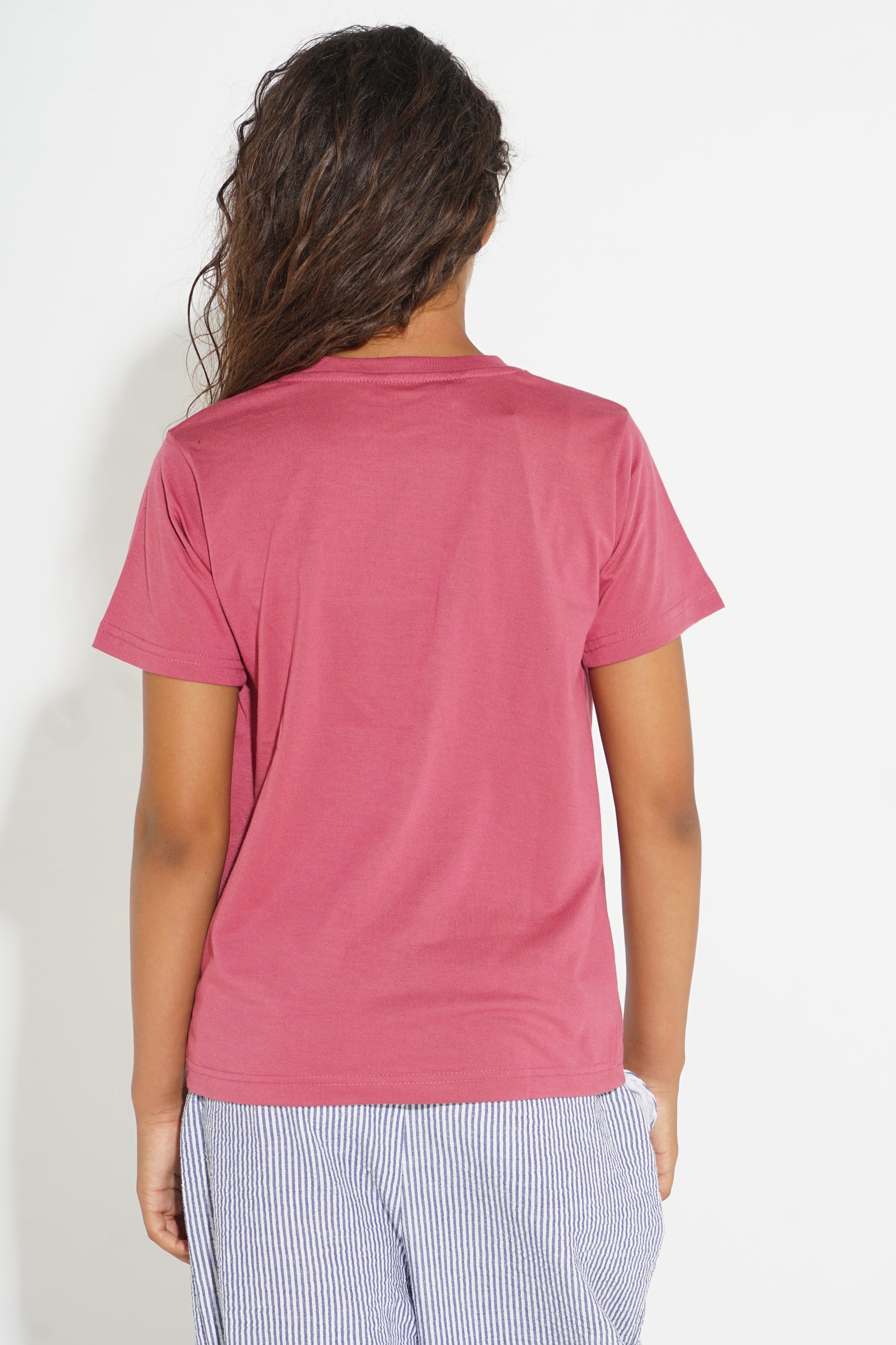 A woman viewed from the back wearing a short-sleeve crewneck t-shirt in a dusty rose pink color, paired with light blue and white striped pants, against a plain white background.