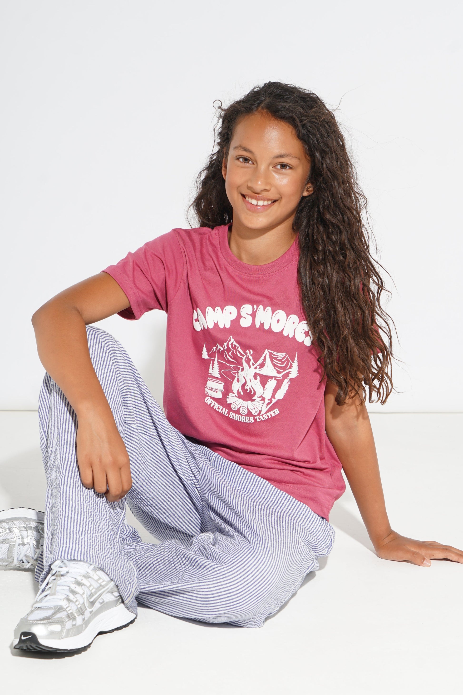 A smiling girl with long curly brown hair sits on a white floor against a white wall, wearing a pink graphic t-shirt that reads 'CAMP SMORE' with a campfire and tent design, light blue and white striped pants, and white sneakers.