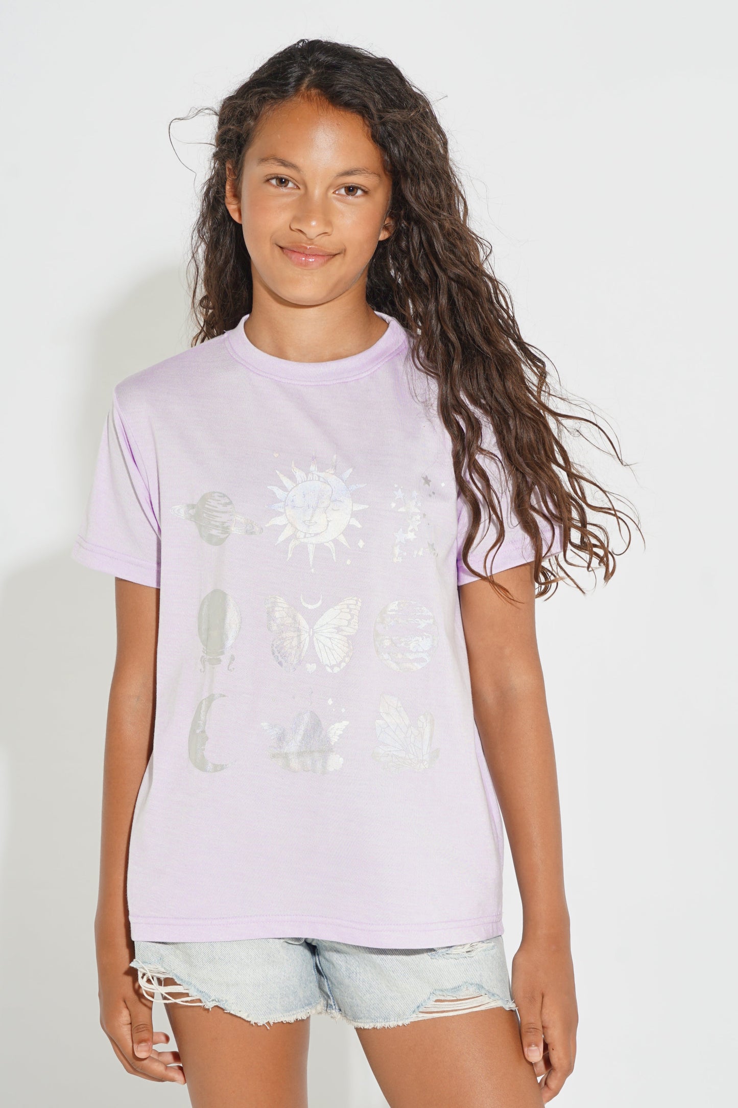 A young girl with long, wavy brown hair wearing a light purple t-shirt with a white celestial graphic print, paired with light-wash denim shorts, standing against a plain white background.