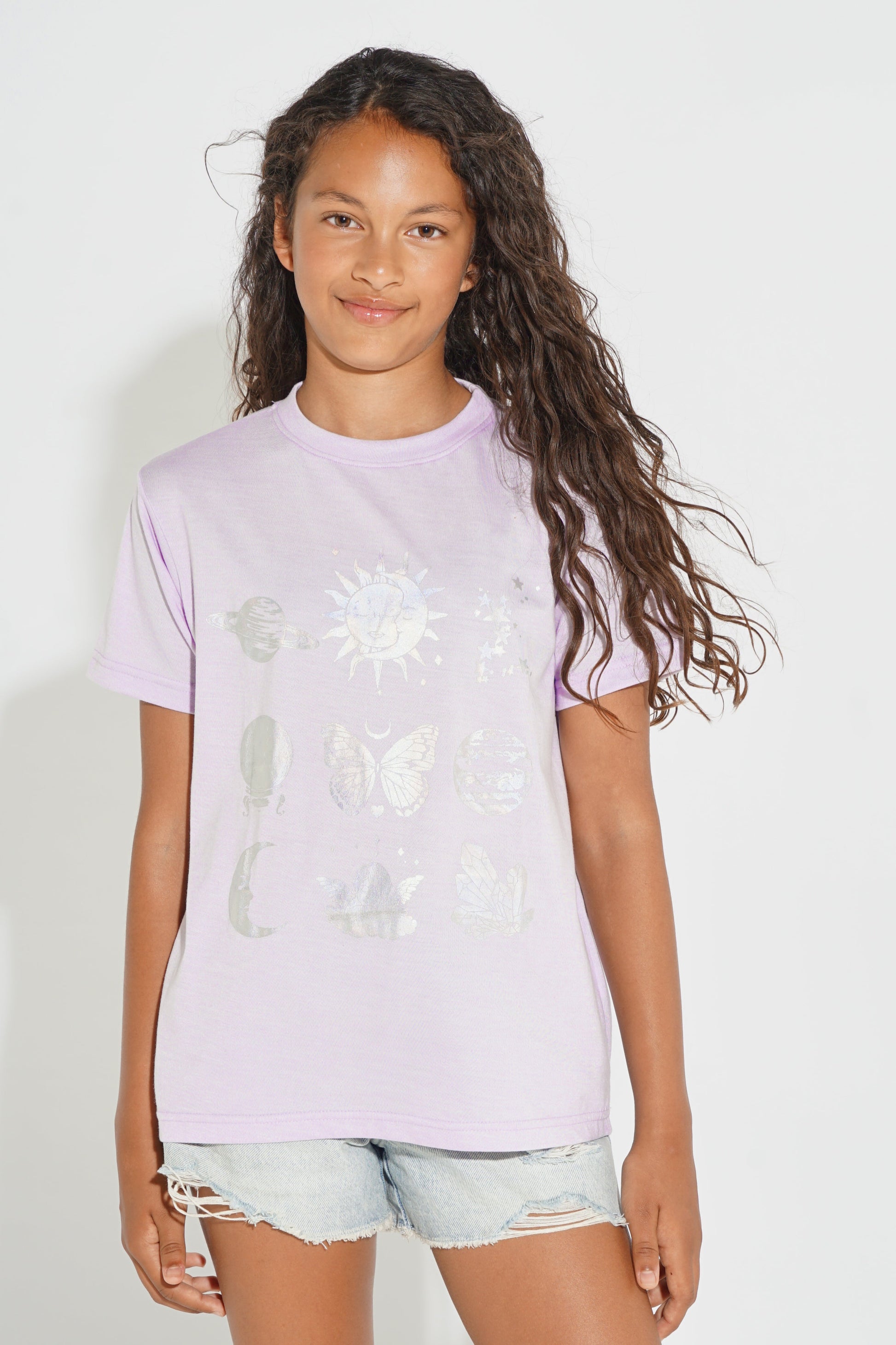 A young girl with long, wavy brown hair wearing a light purple t-shirt with a white celestial graphic print, paired with light-wash denim shorts, standing against a plain white background.