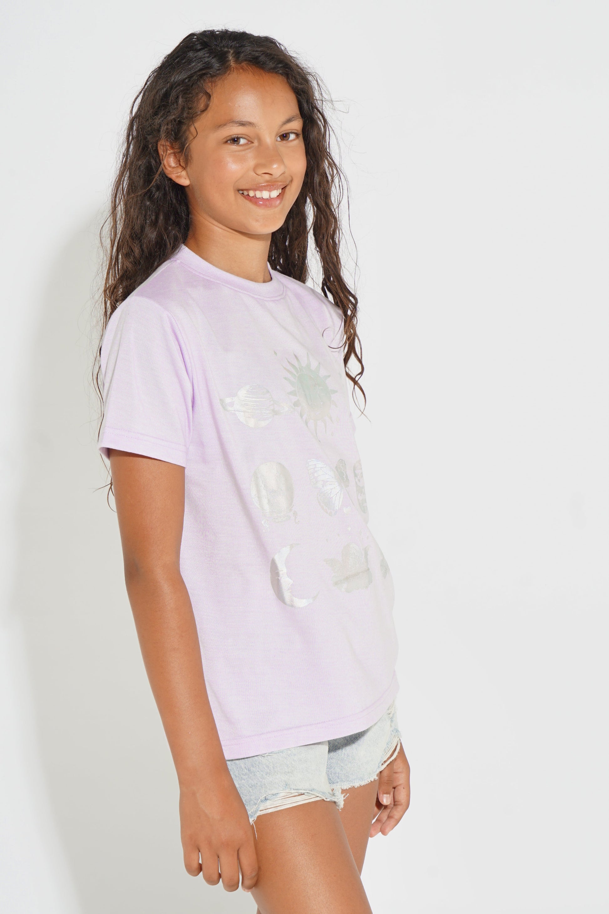 A young girl with long dark curly hair smiling in a side profile, wearing a light lavender short-sleeve t-shirt with a distressed graphic print and light denim shorts, set against a plain white studio background.