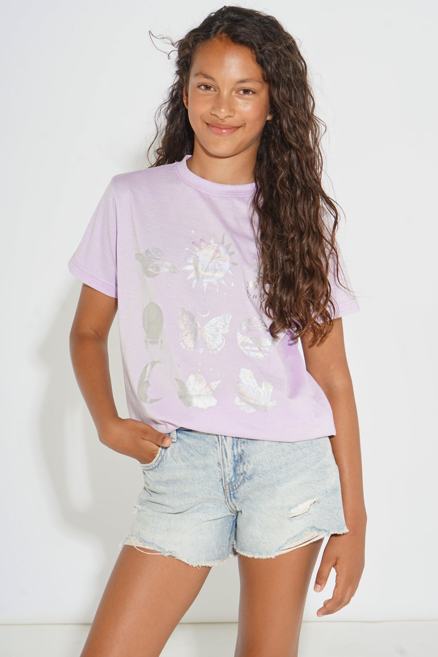 A young girl with long, curly brown hair wearing a light purple t-shirt with white butterfly and floral graphics, paired with light-wash denim shorts, standing against a white studio background.