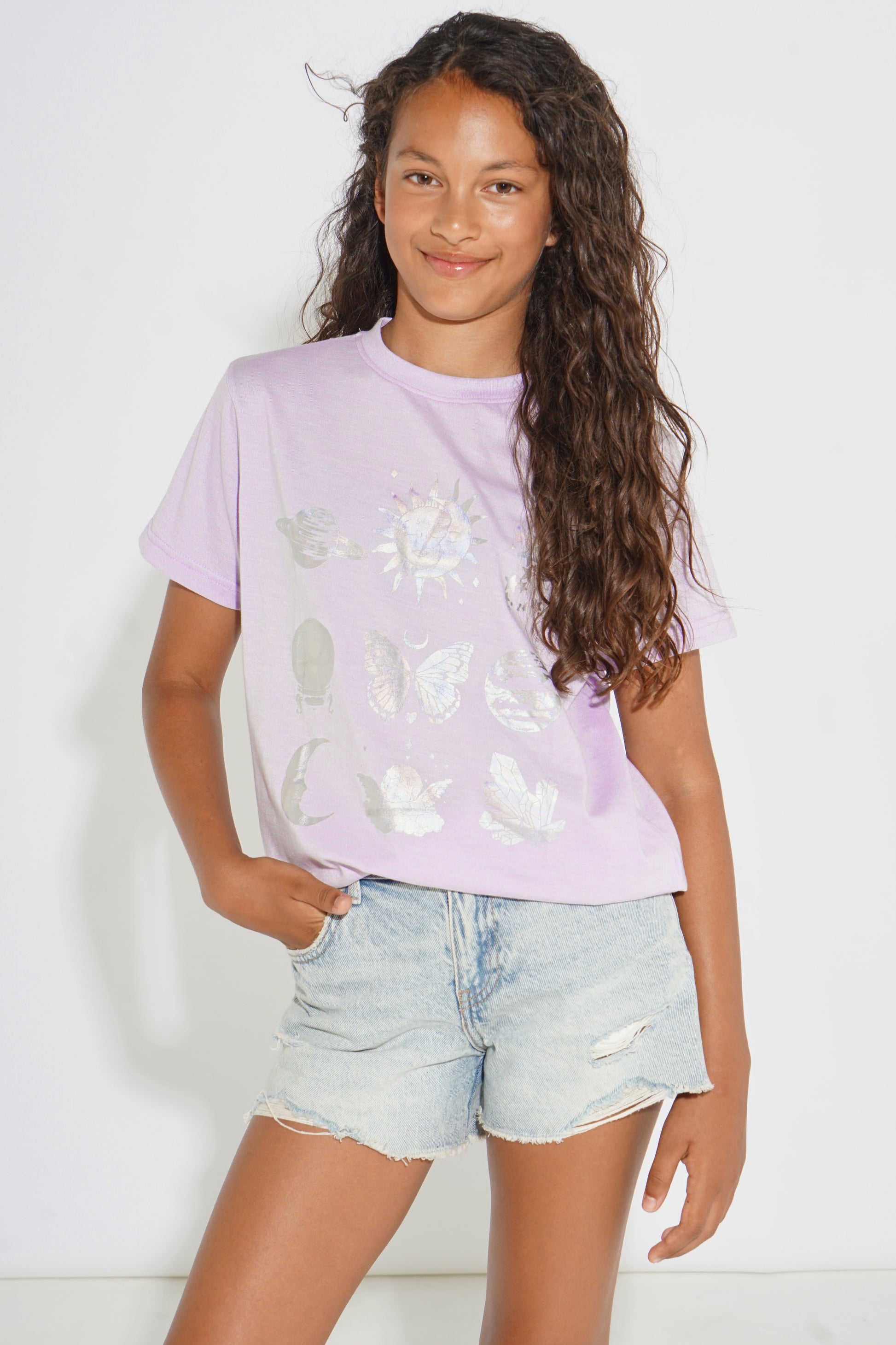A young girl with long, curly brown hair wearing a light purple t-shirt with white butterfly and floral graphics, paired with light-wash denim shorts, standing against a white studio background.