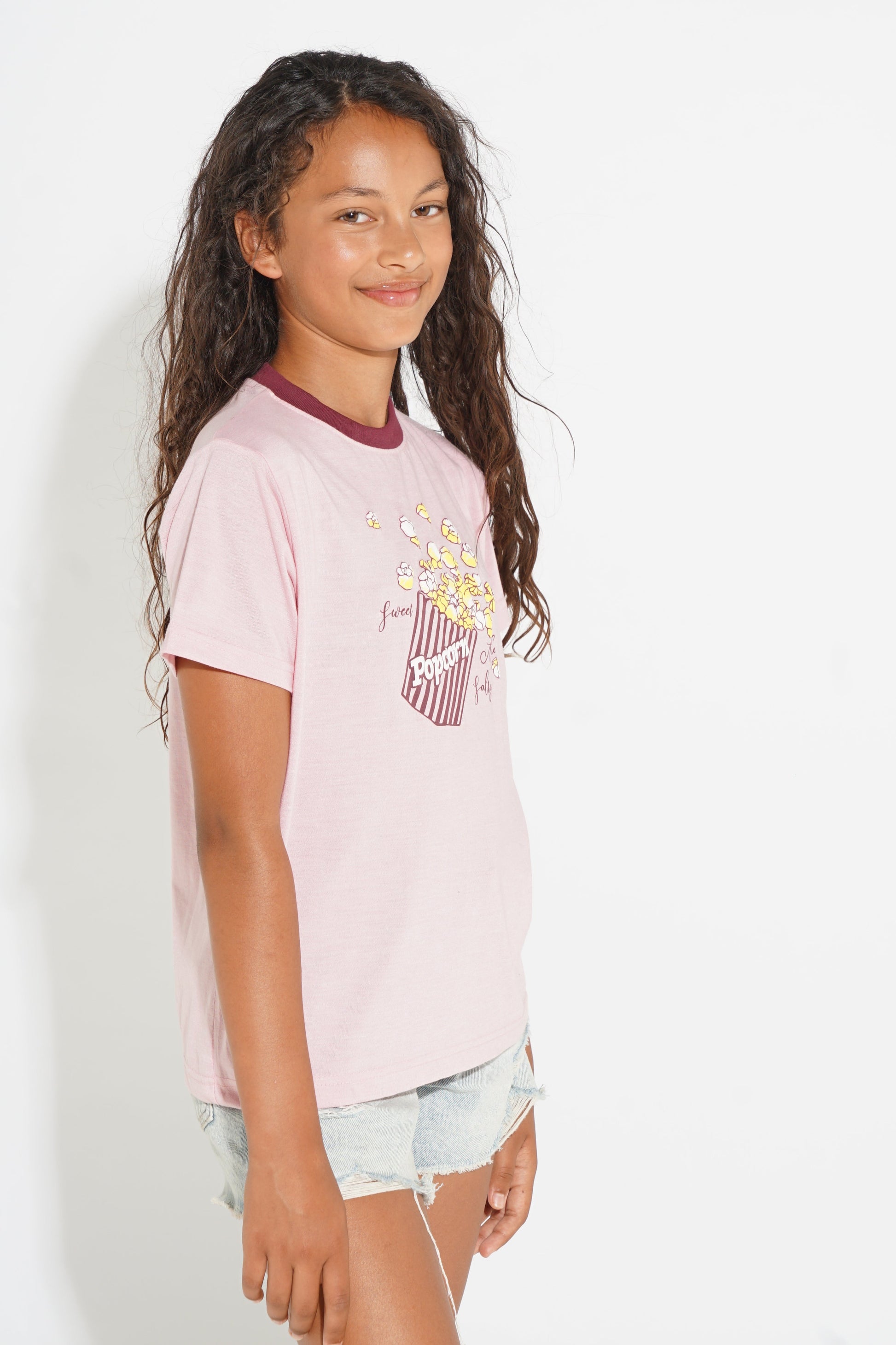 A young girl with long, wavy brown hair is shown from the waist up, wearing a light pink graphic t-shirt with a popcorn bucket design and light-wash denim shorts, set against a plain white studio background.