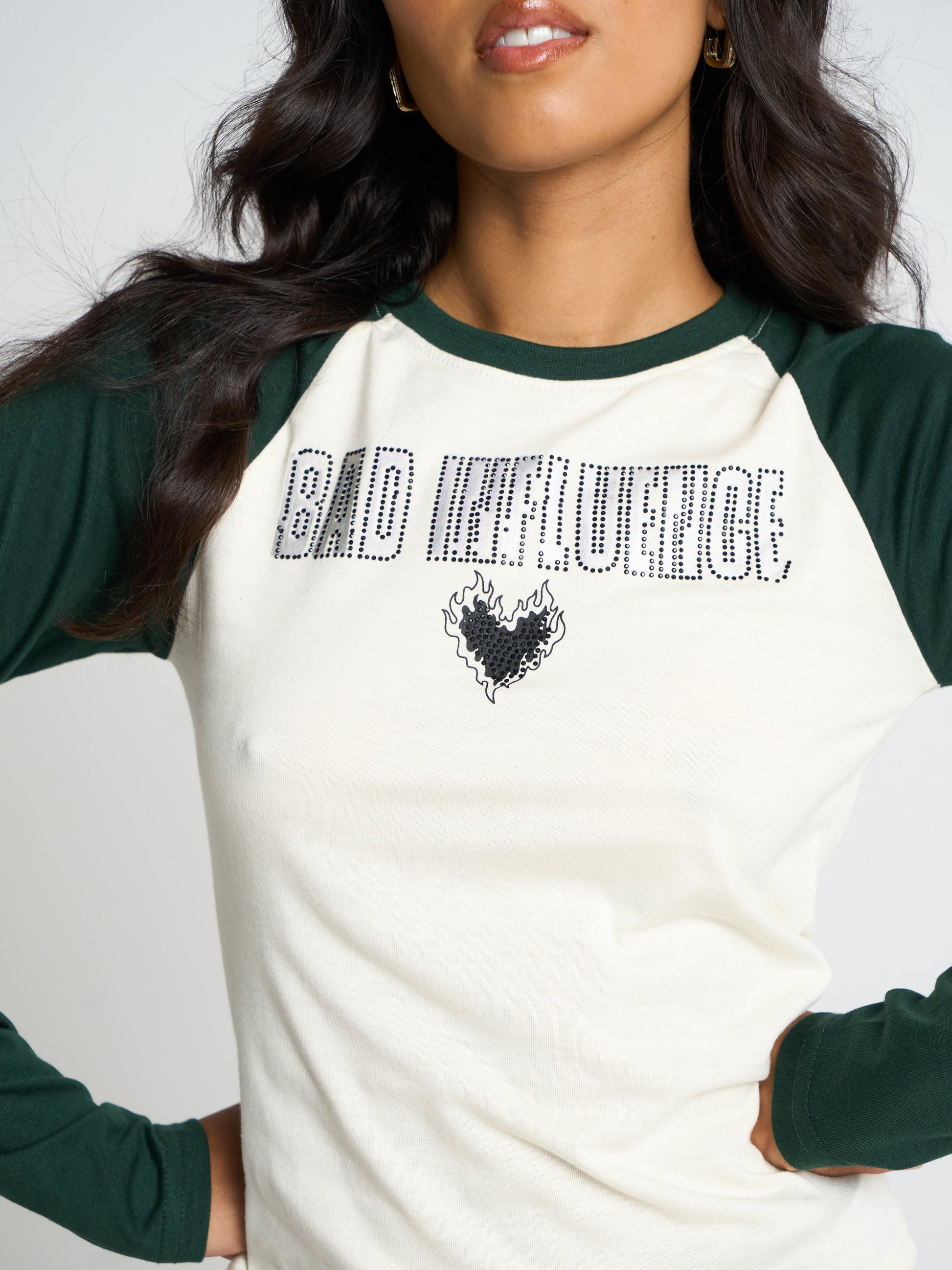 A woman wearing a raglan-style long-sleeve shirt with an off-white body and dark green sleeves. The shirt features rhinestone text reading 'BAD INFLUENCE' and a black heart graphic on the chest.