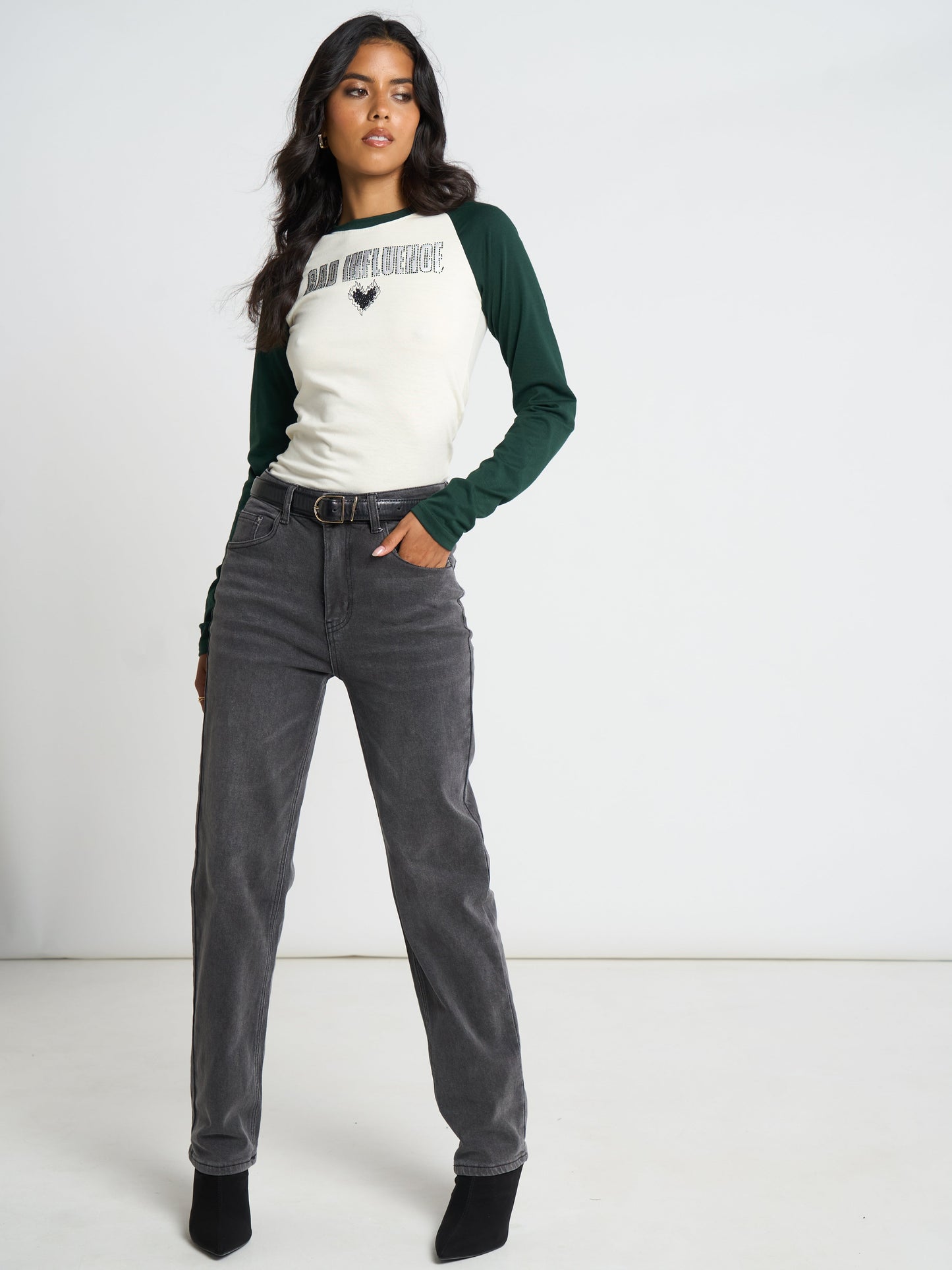 A woman modeling a raglan-style long-sleeve top with a cream front and dark green sleeves, paired with dark gray jeans and black ankle boots, against a plain white studio background.