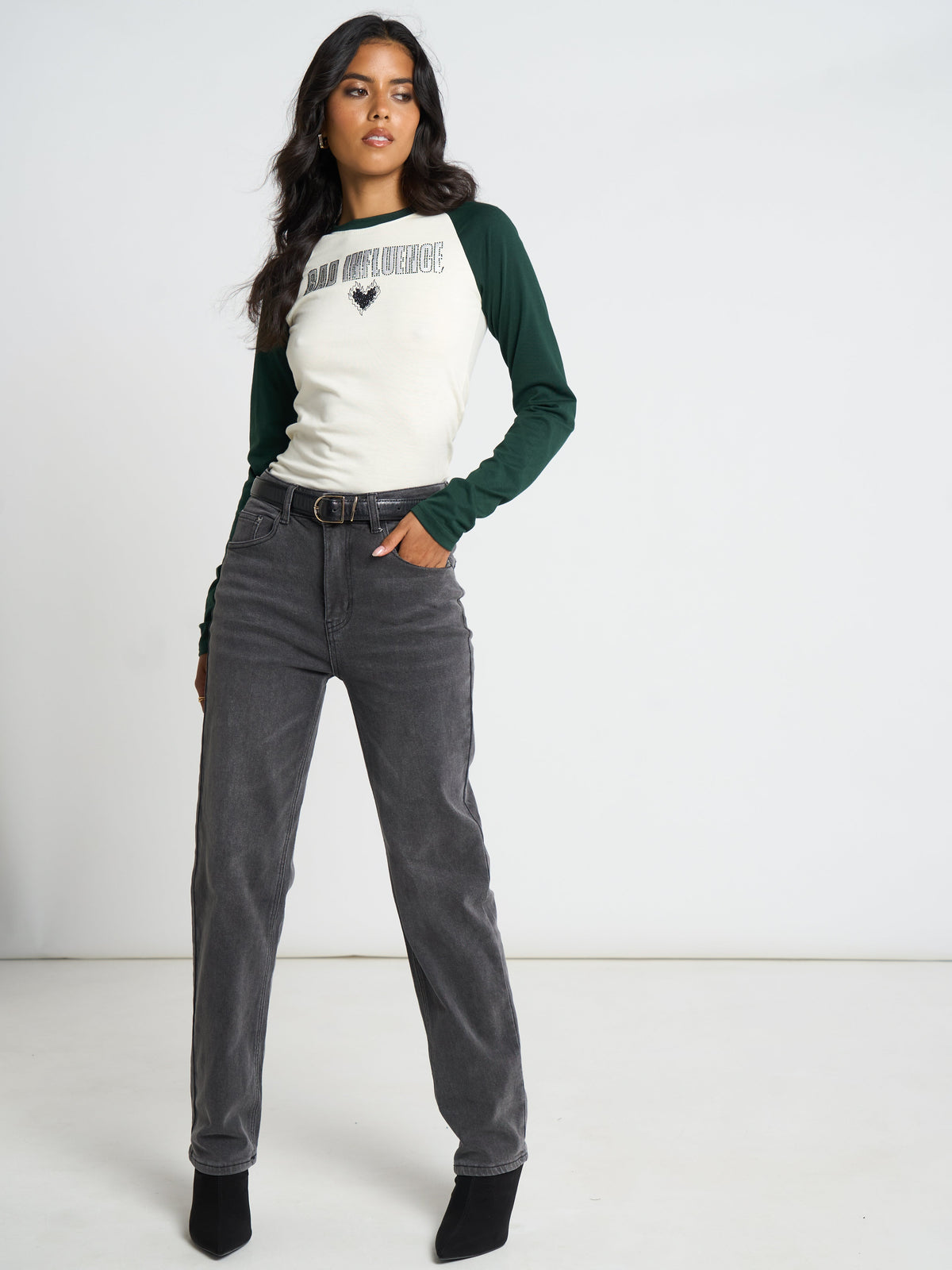 A woman modeling a raglan-style long-sleeve top with a cream front and dark green sleeves, paired with dark gray jeans and black ankle boots, against a plain white studio background.