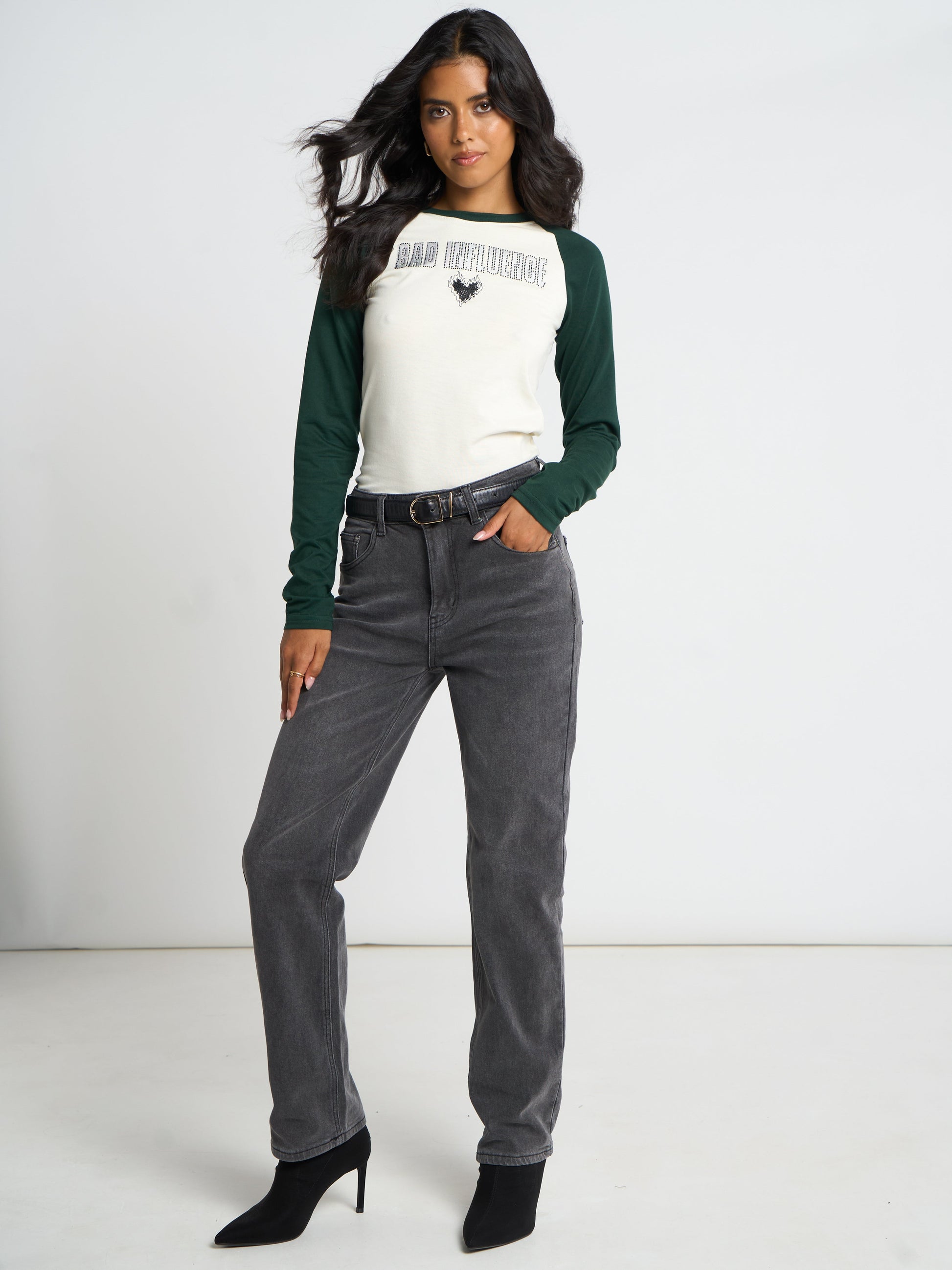 A woman modeling a white and dark green raglan-style long-sleeve shirt with 'SAN DIEGO STATE' and a black duck logo on the chest, paired with gray jeans and black ankle boots against a plain white studio background.