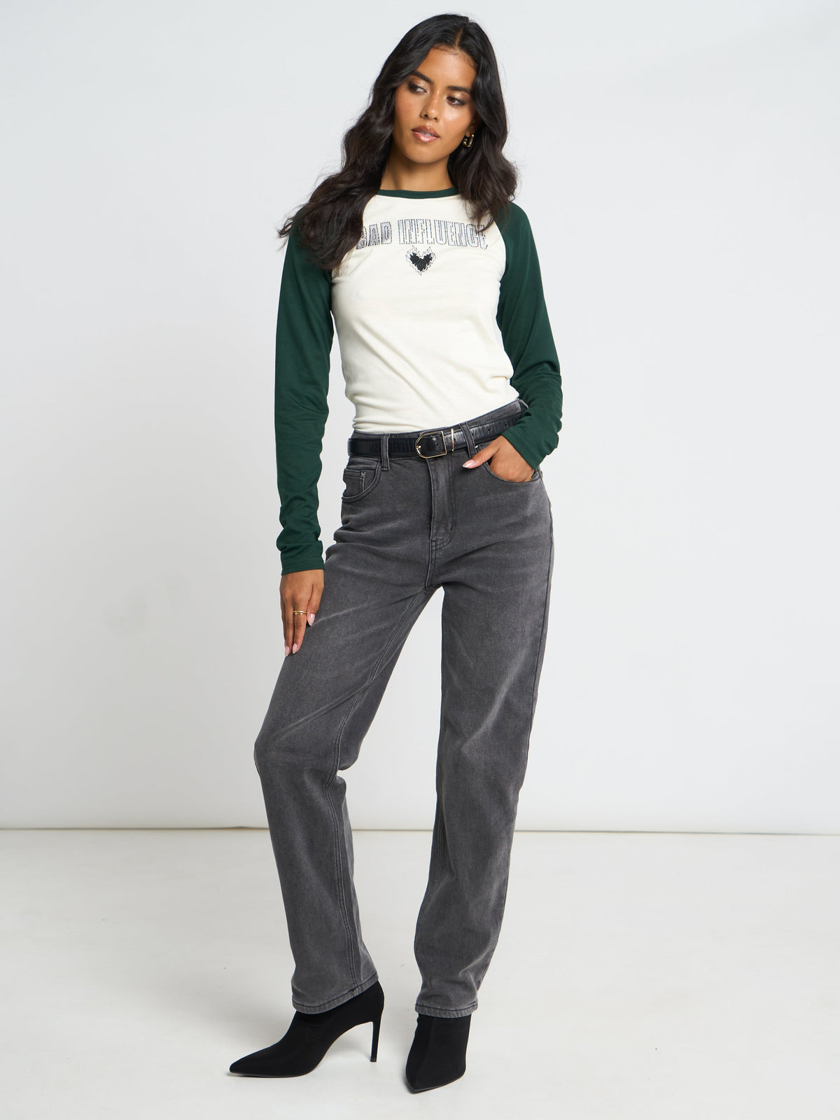 A woman modeling a white and dark green raglan long-sleeve shirt with 'PINE RIDGE' and a heart graphic on the chest, paired with dark gray jeans and black heels, against a plain white studio background.