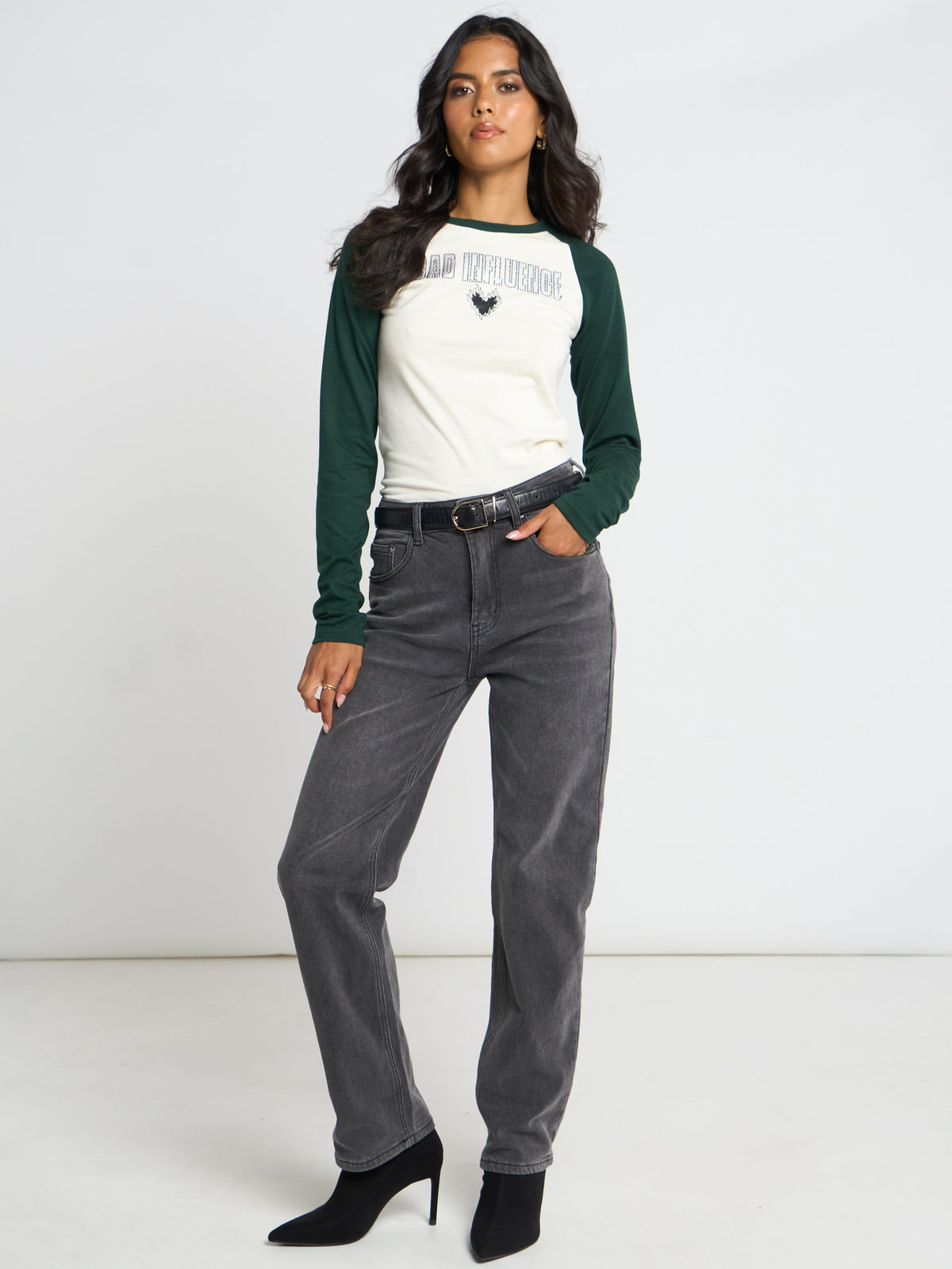 A woman modeling a white and dark green raglan long-sleeve shirt with 'AN TOURNING' text and a rooster graphic on the chest, paired with dark gray jeans and black heels against a white studio background.