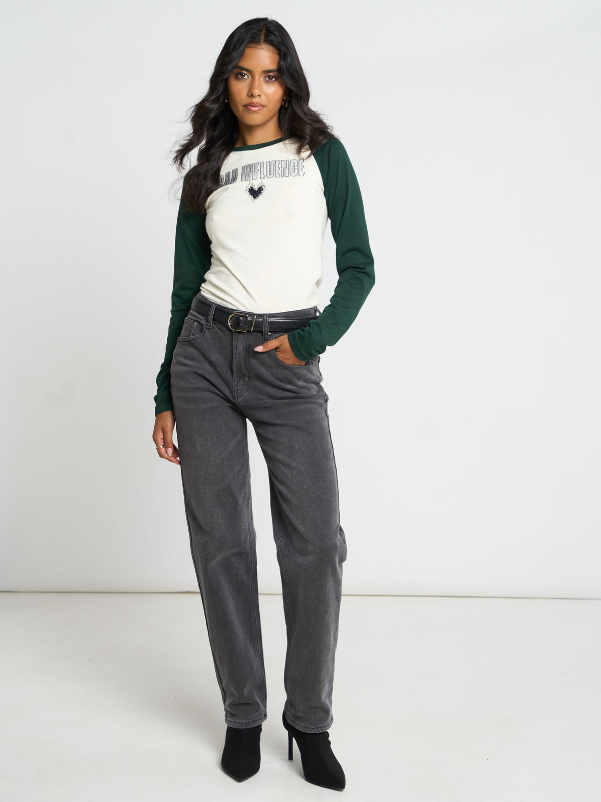 A woman modeling a white and dark green raglan long-sleeve t-shirt with a small black heart graphic on the chest, paired with dark gray jeans and black ankle boots, against a plain white studio background.
