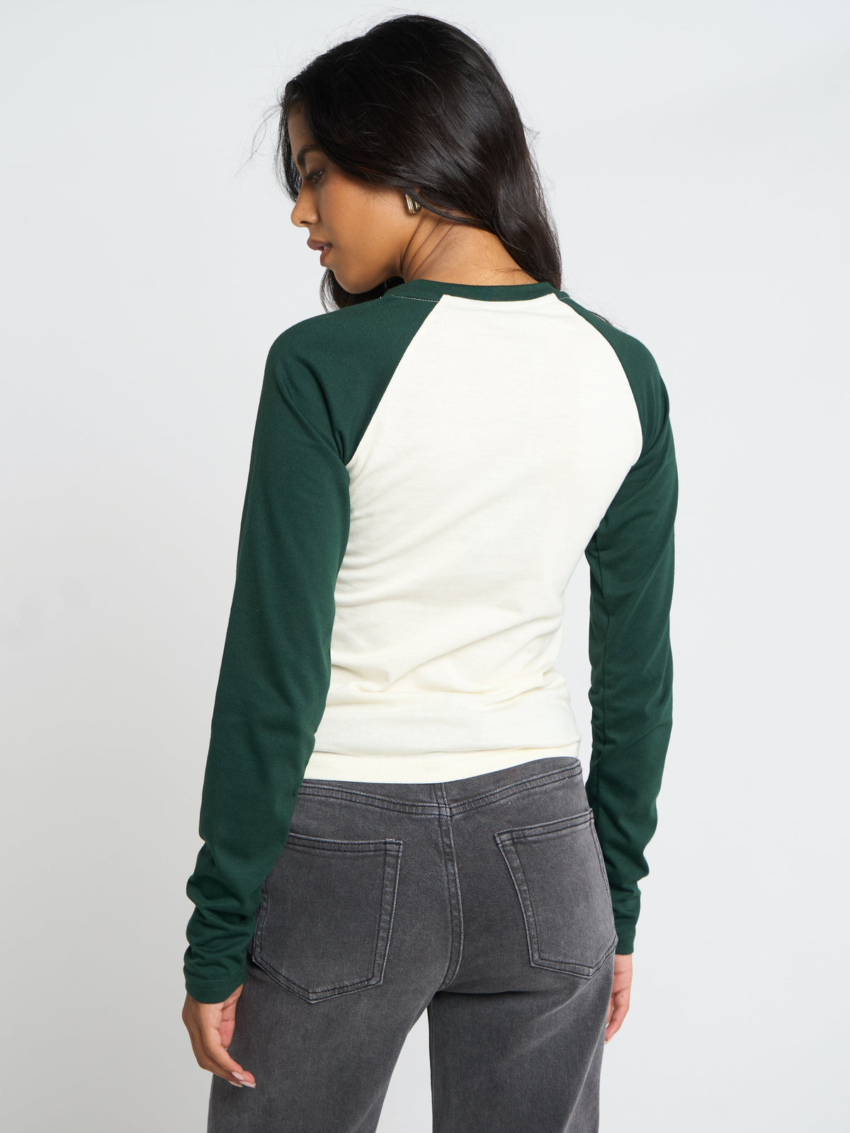 A woman shown from the back wearing a long-sleeve raglan shirt with a cream body and dark green sleeves, paired with dark gray jeans, against a light gray studio background.