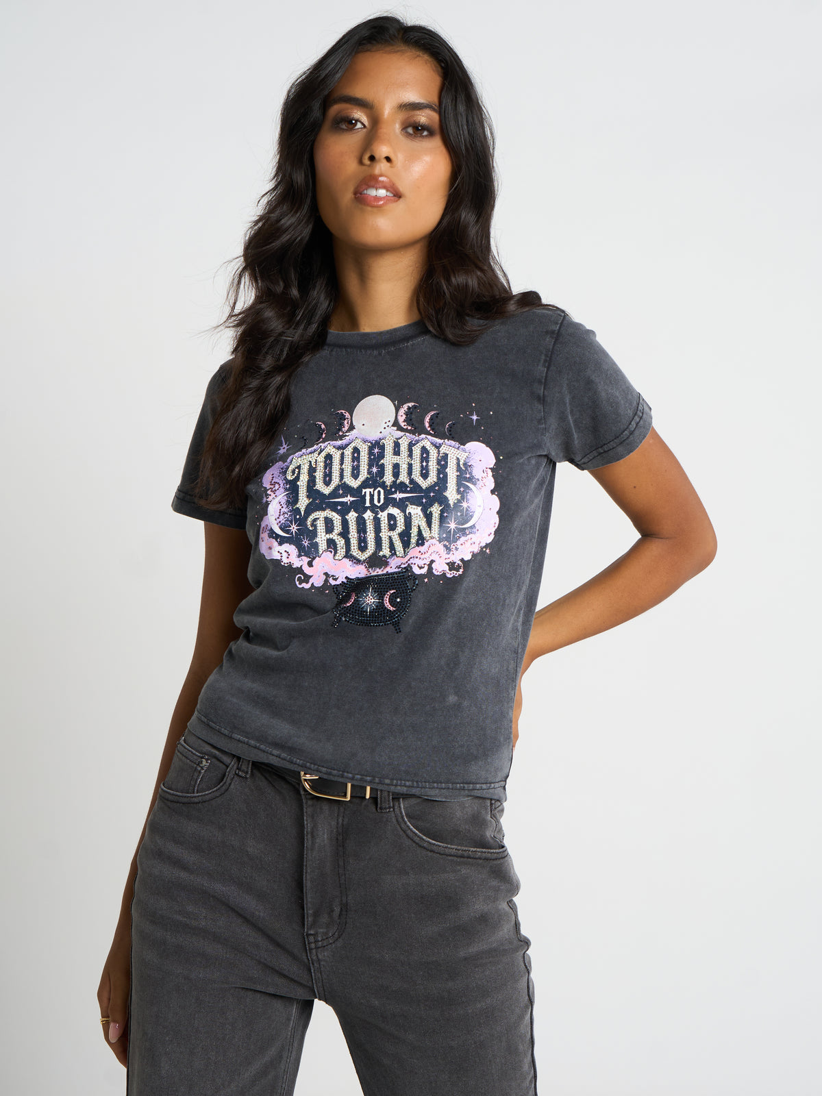 A woman modeling a dark gray acid-wash graphic t-shirt with 'Too Hot To Burn' in a pink and white celestial design, paired with dark gray jeans against a light gray studio background.