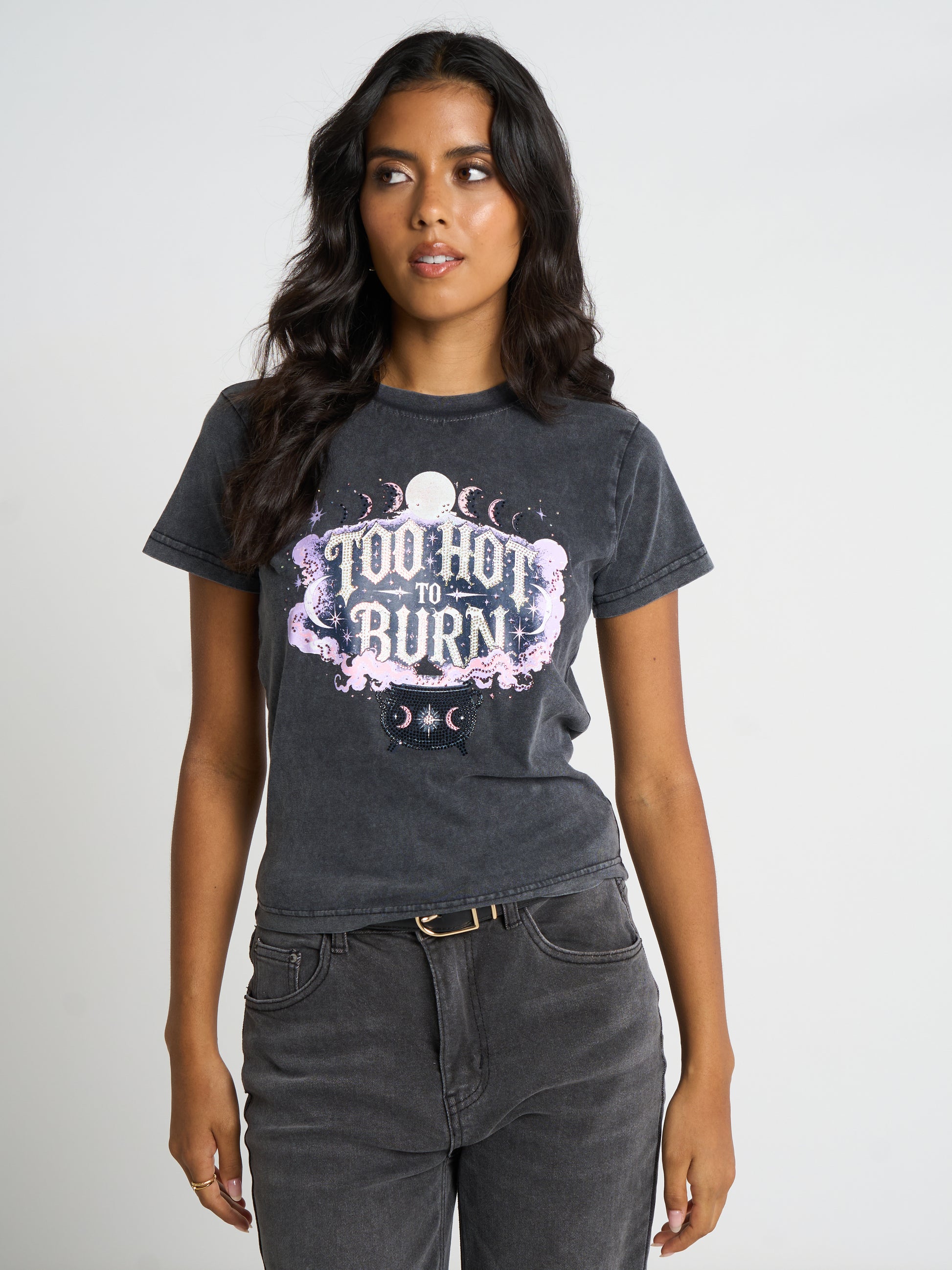 A woman modeling a dark gray acid-wash graphic t-shirt with a pink and white 'Too Ho to Burn' moon design, paired with dark gray jeans against a light gray studio background.