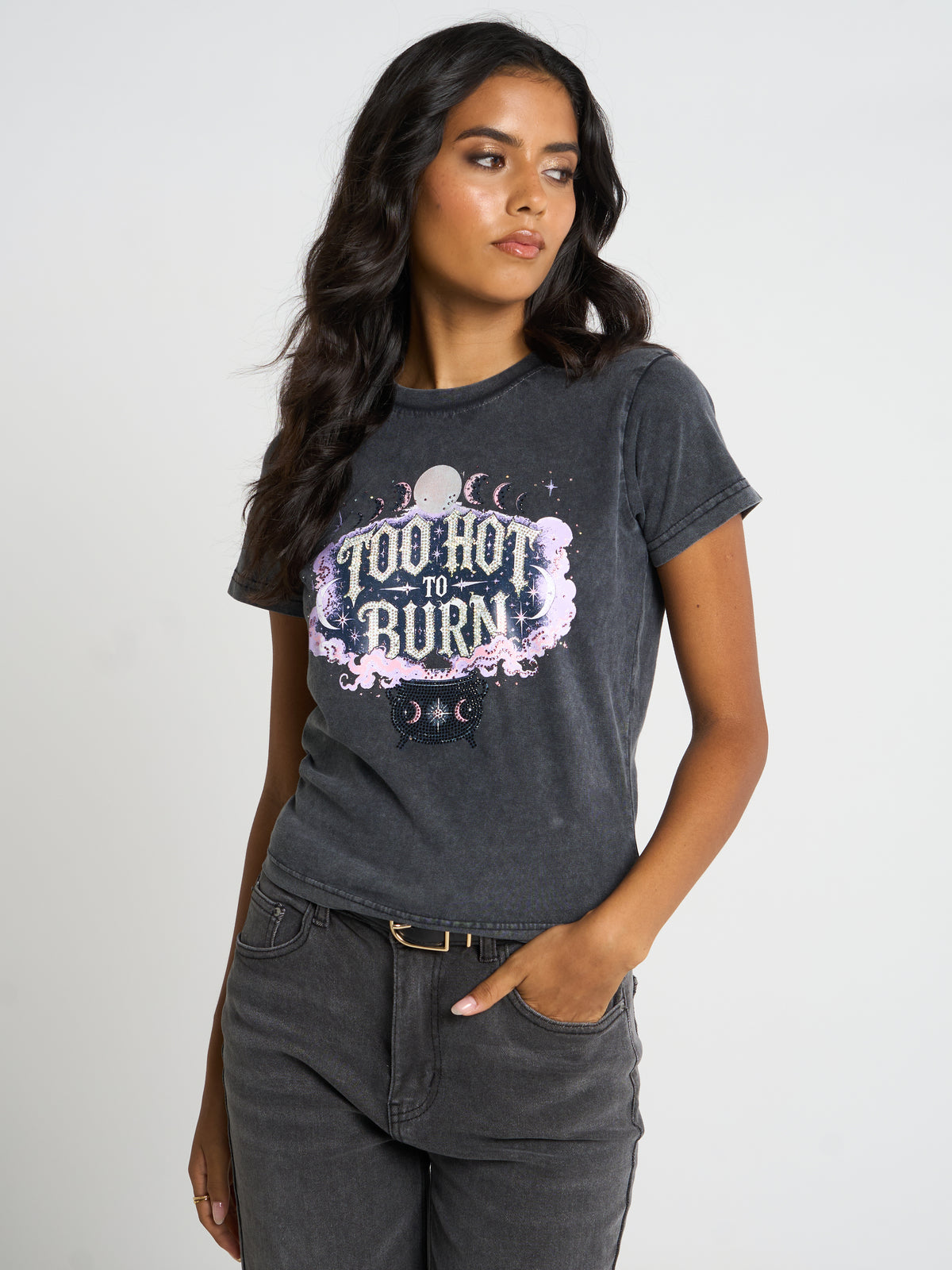 A woman modeling a dark gray acid-wash graphic t-shirt with 'Too Hot to Burn' in a pink and purple celestial design, paired with dark gray jeans against a light gray studio background.