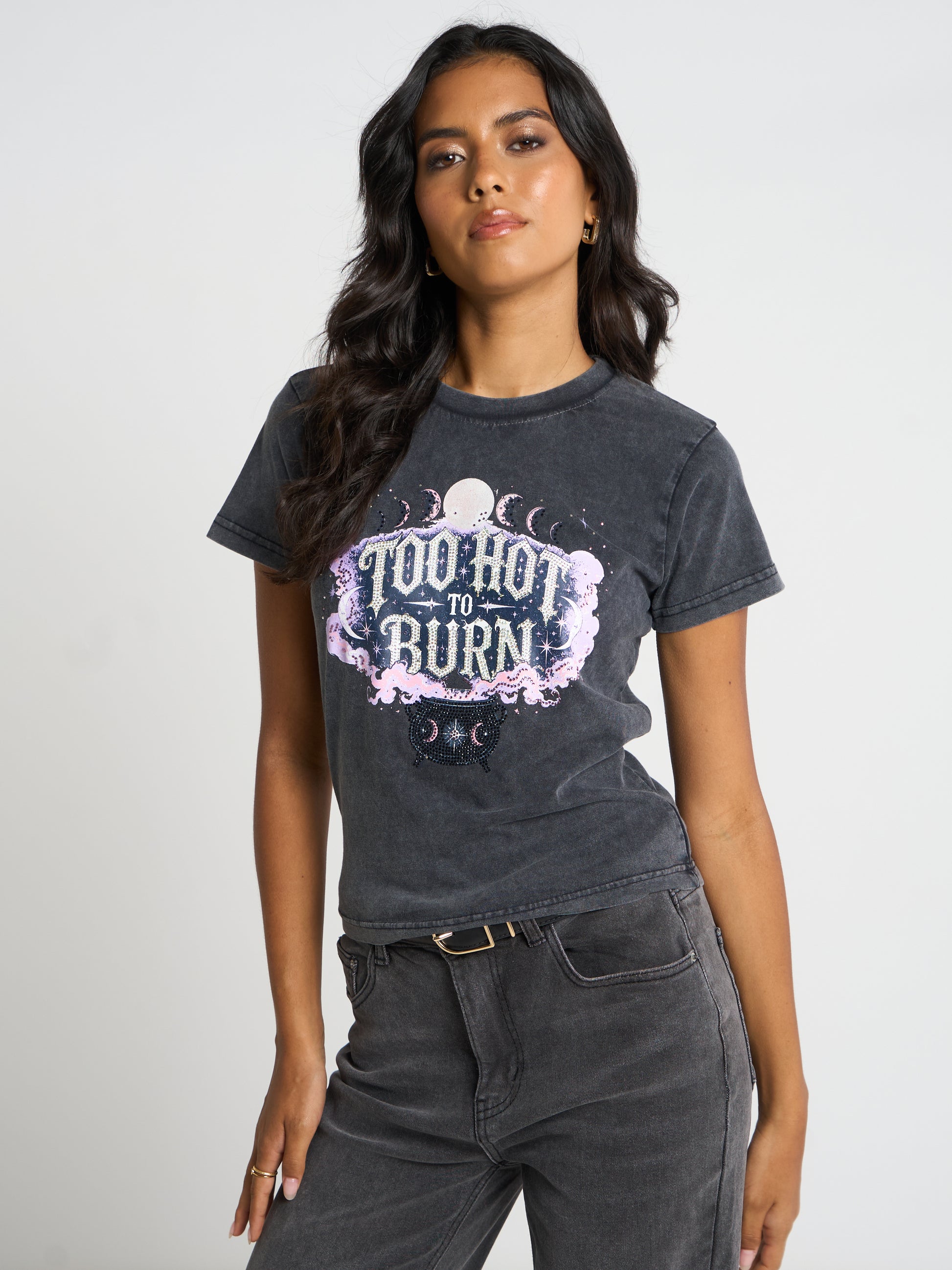 A woman modeling a dark gray washed graphic t-shirt with a 'Too Hot to Burn' moon and cat design, paired with dark gray jeans against a light gray studio background.