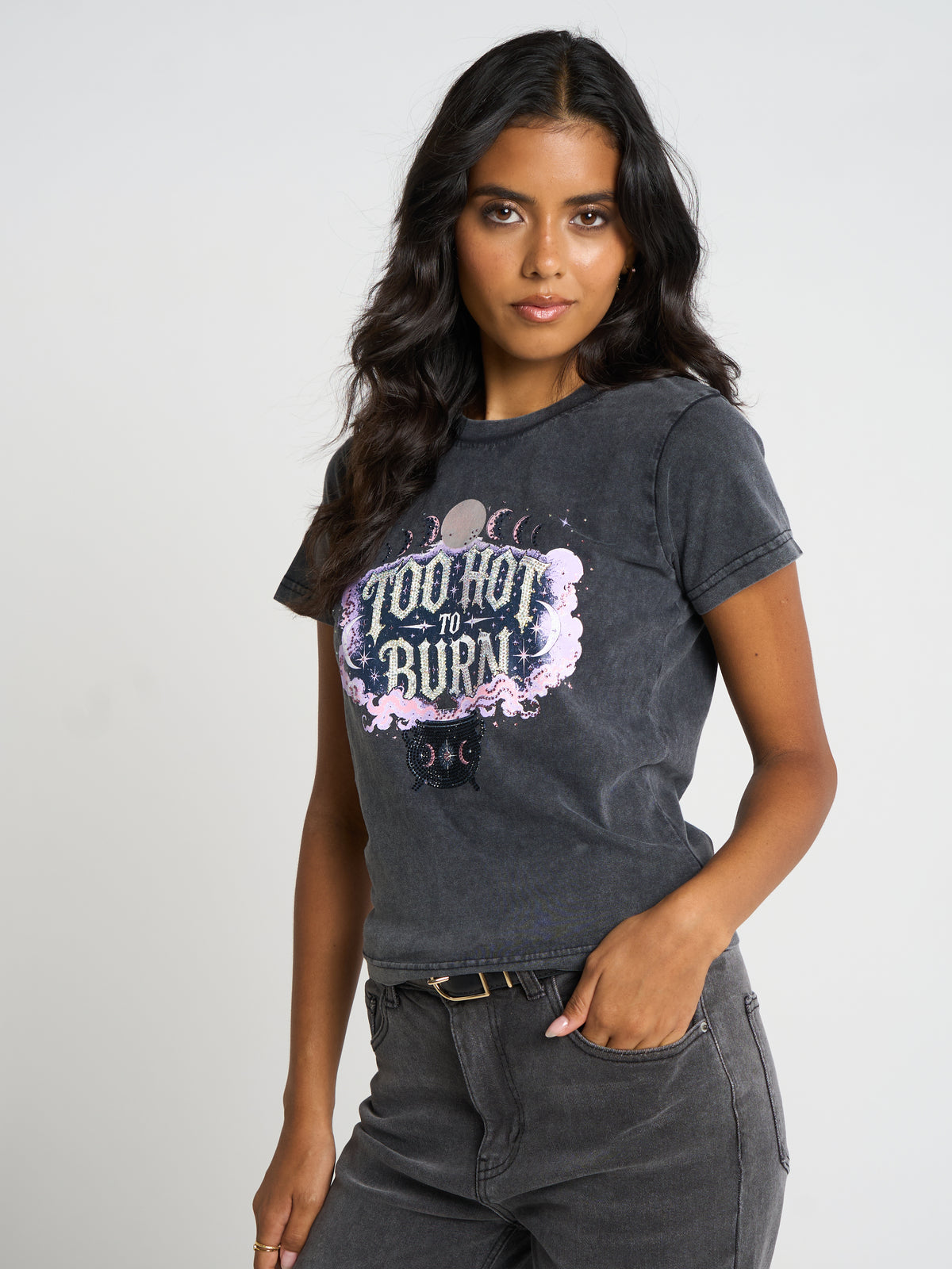 A woman modeling a dark gray acid-wash graphic t-shirt with a 'Too Hot to Burn' design, paired with dark gray jeans, against a light gray studio background.