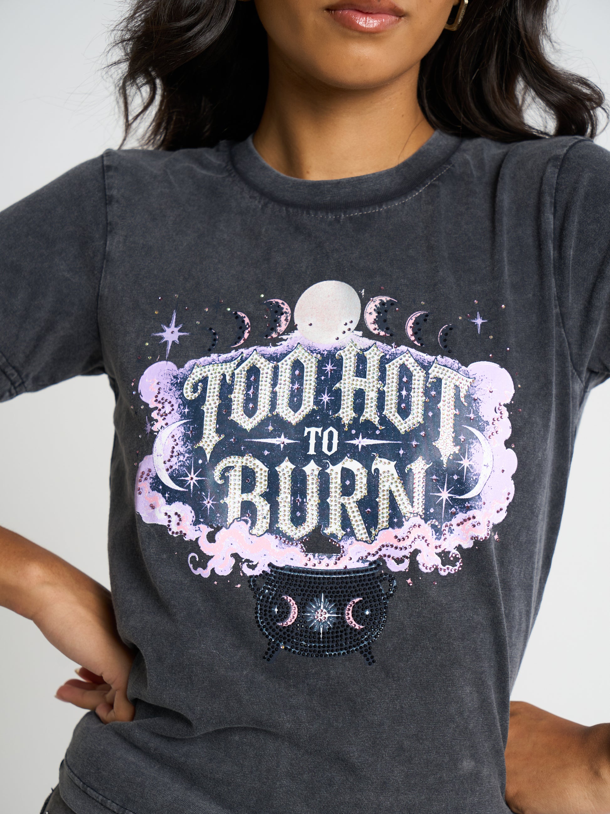 A woman wearing a dark gray acid-wash graphic t-shirt with a celestial 'Too Hot to Burn' design featuring a moon, stars, and a cauldron.