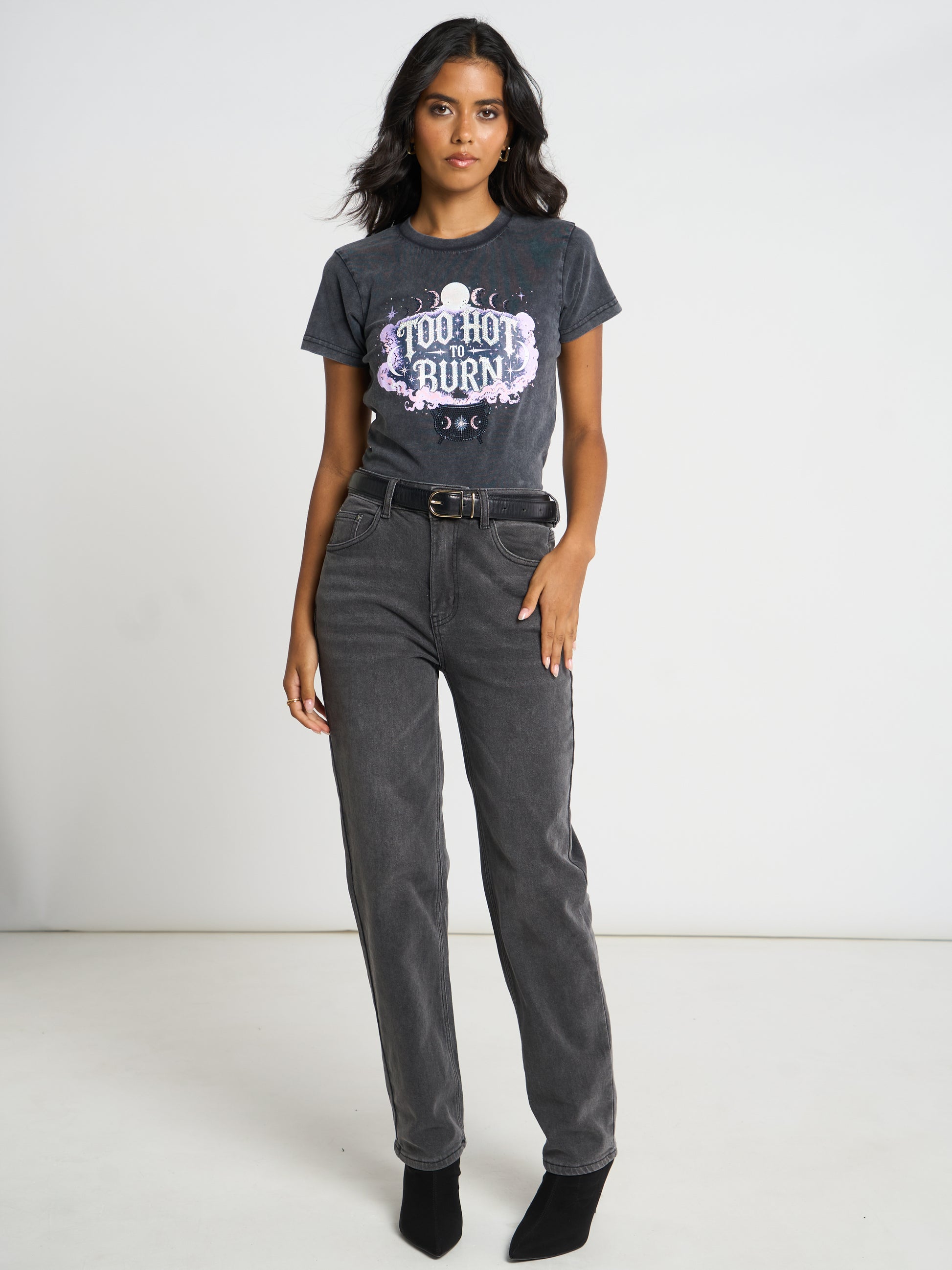 A woman modeling a dark gray acid-wash graphic t-shirt with 'YOU HOT BURN' in white and purple, paired with dark gray jeans, a black belt, and black ankle boots against a white studio background.
