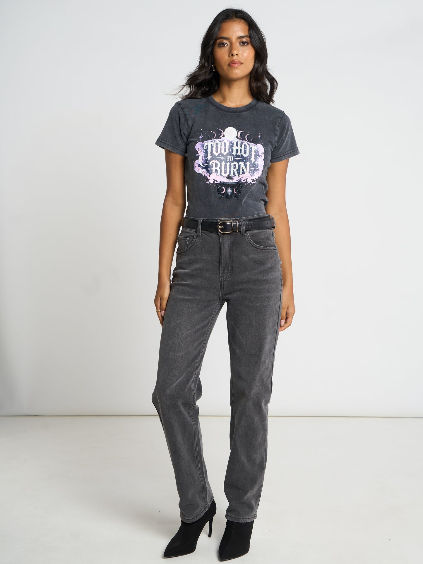 A woman modeling a washed black graphic t-shirt with 'Too Hot to Burn' text, paired with dark gray jeans and black ankle boots, against a plain white studio background.