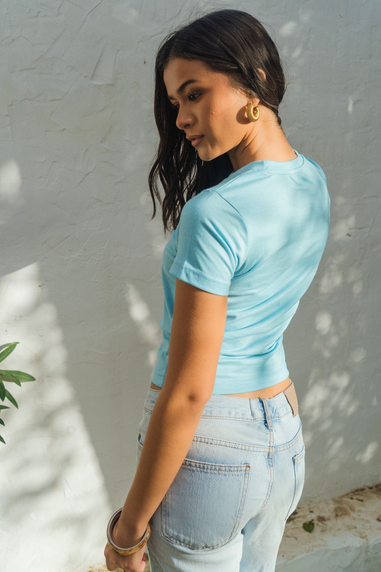 Woman wearing a light blue top and jeans against a white wall.