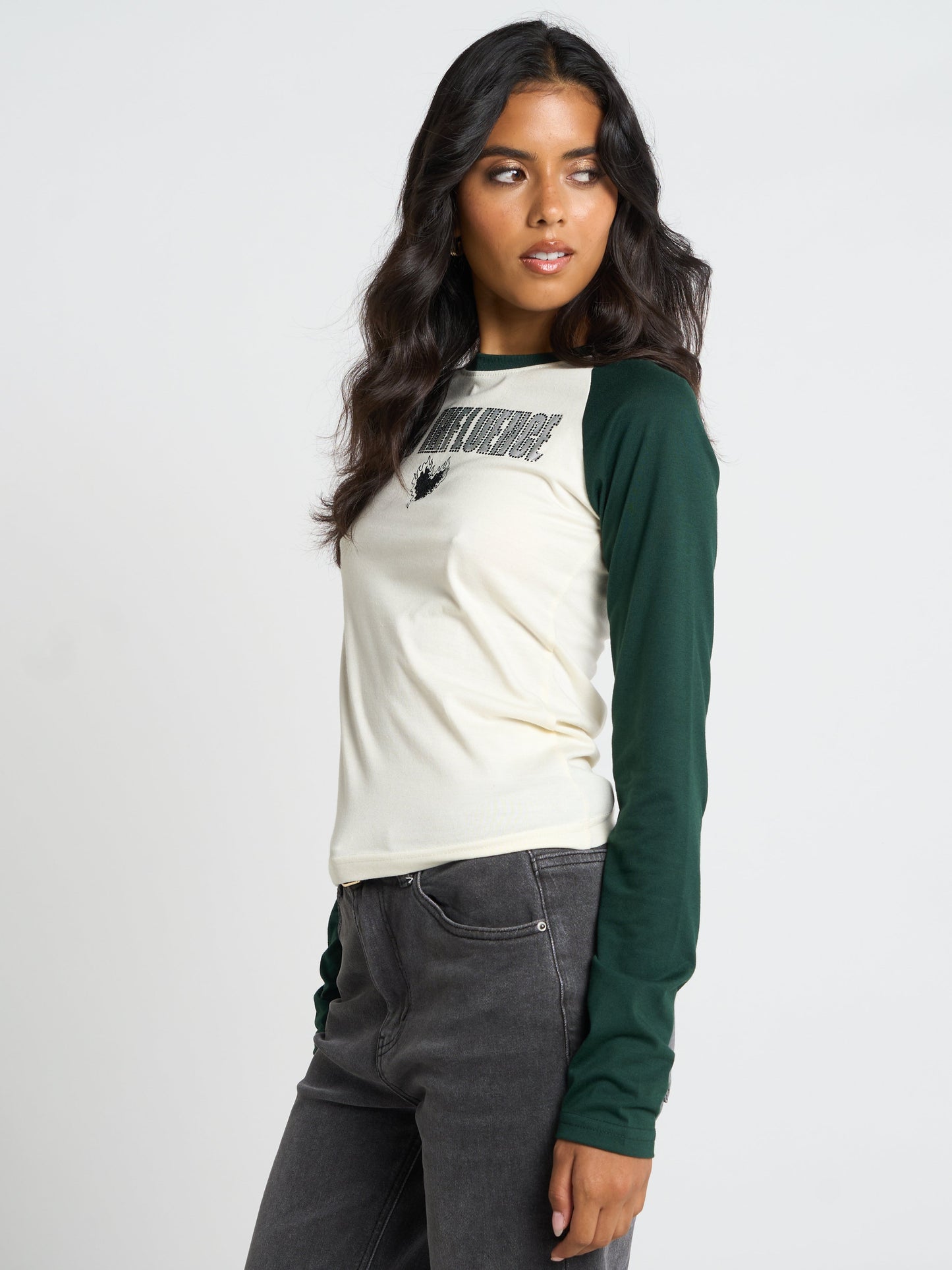 A woman modeling a long-sleeve raglan t-shirt with an off-white body and dark green sleeves, featuring a black graphic print on the chest, paired with dark gray jeans against a light gray studio background.
