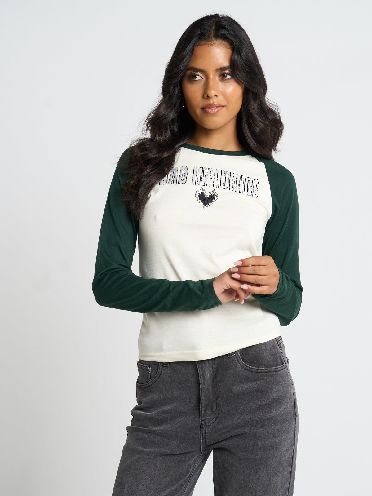 A woman modeling a long-sleeve raglan t-shirt with an off-white body and dark green sleeves. The shirt features the text 'BAD INFLUENCE' in a distressed stencil font across the chest, above a black rooster graphic.