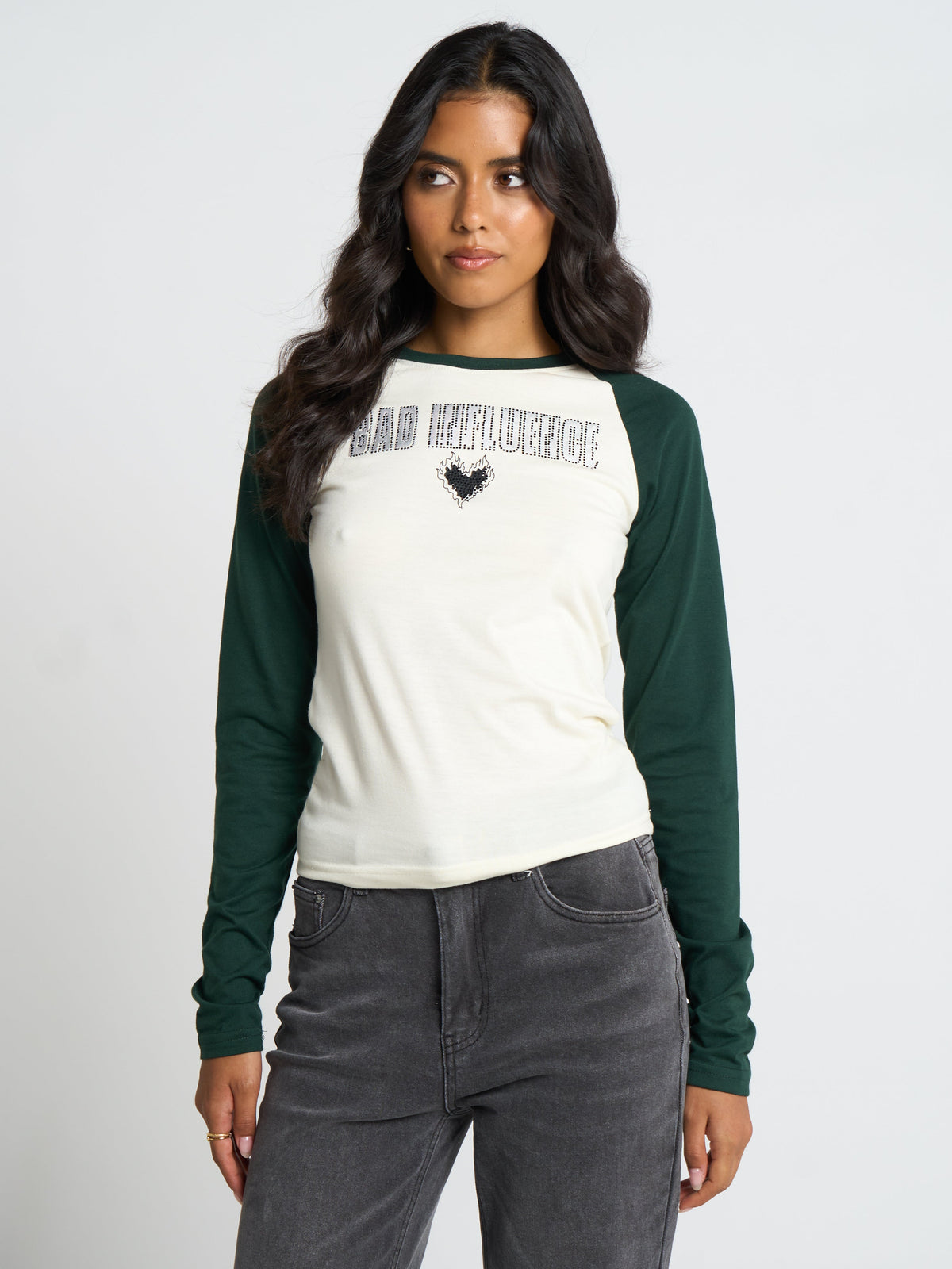 A woman modeling a long-sleeve raglan t-shirt with an off-white body and dark green sleeves, featuring a graphic print on the chest that reads 'BAD INFLUENCE' with a black rooster silhouette.