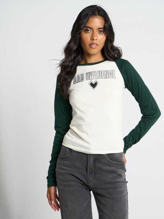A woman modeling a raglan-style long-sleeve shirt with an off-white body and dark green sleeves. The shirt features the text 'BAD INFLUENCE' in a distressed black font with a small black heart graphic below it.