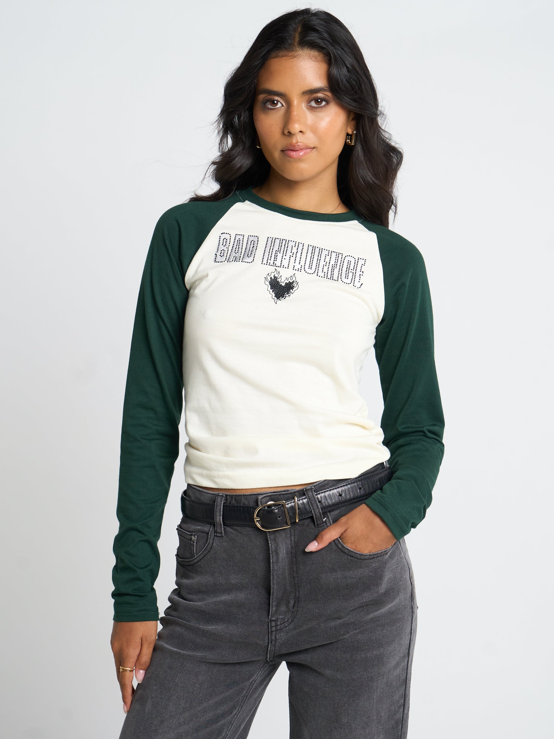 A woman modeling a long-sleeve raglan baseball tee with a cream body and dark green sleeves, featuring 'BAD INFLUENCE' text and a rooster graphic on the chest, paired with dark gray jeans and a black belt against a light gray studio background.