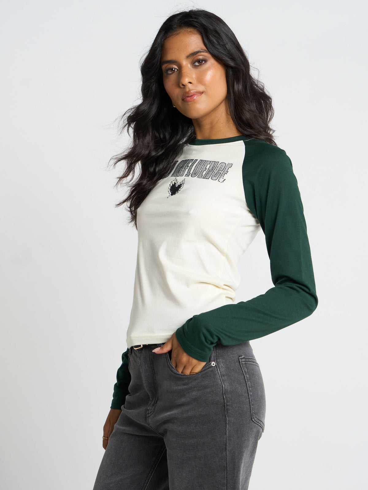 A woman modeling a long-sleeve raglan baseball tee with an off-white body and dark green sleeves, featuring a black graphic print on the chest, paired with dark gray jeans against a plain light gray background.