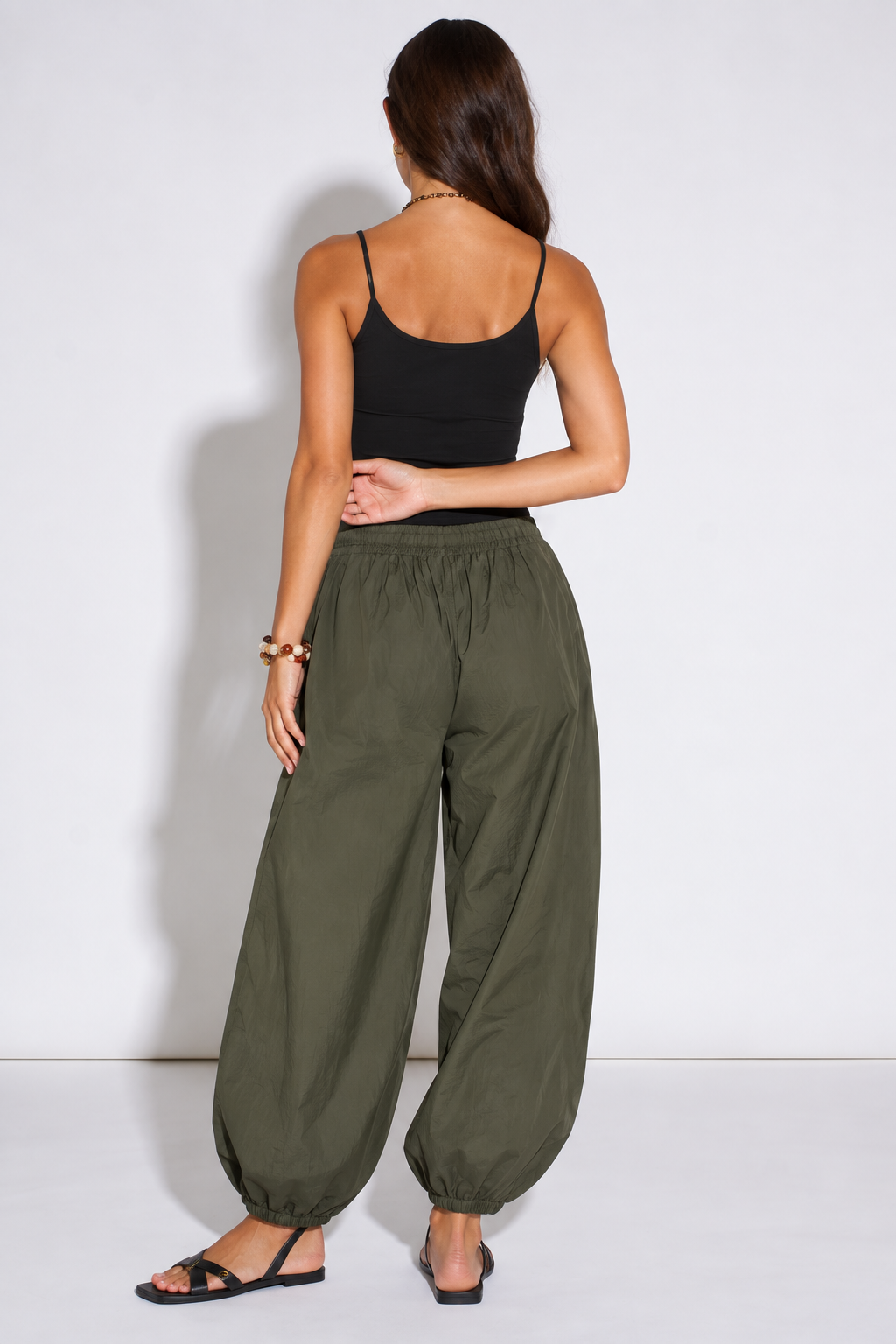 A woman shown from the back wearing loose, wide-leg olive green harem-style pants with an elastic waist and gathered cuffs, paired with a black spaghetti-strap tank top and black sandals, against a plain white studio background.