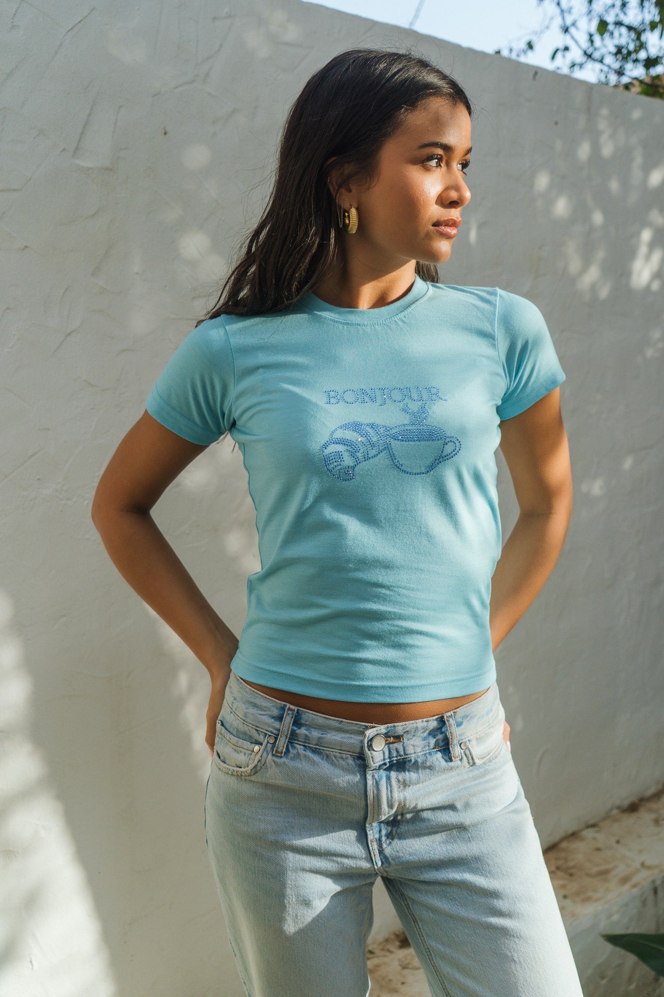 Woman wearing a light blue t-shirt with text and graphics, standing against a white wall.