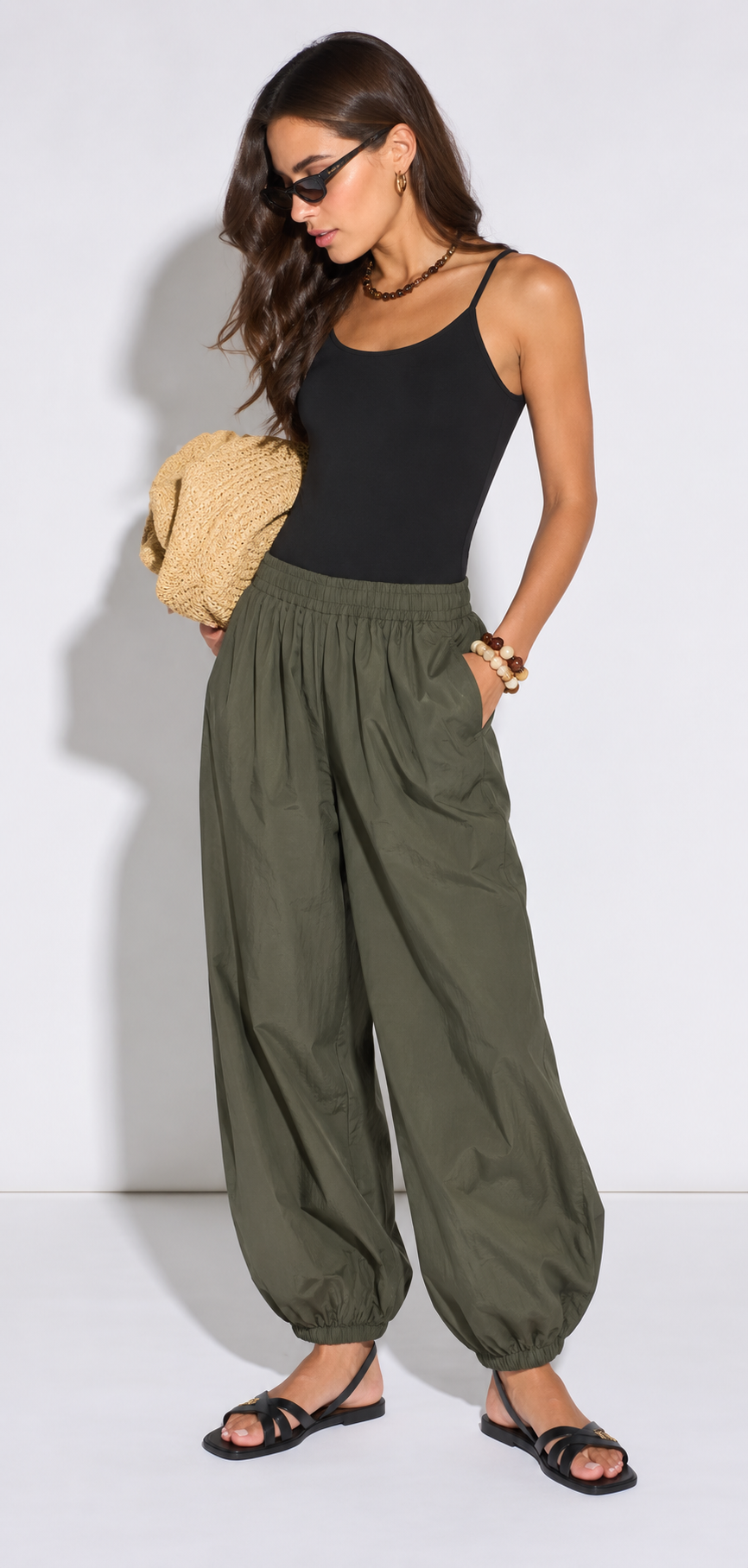 A woman modeling a black sleeveless tank top and olive green wide-leg harem pants, styled with black sandals and a straw clutch against a plain white studio background.