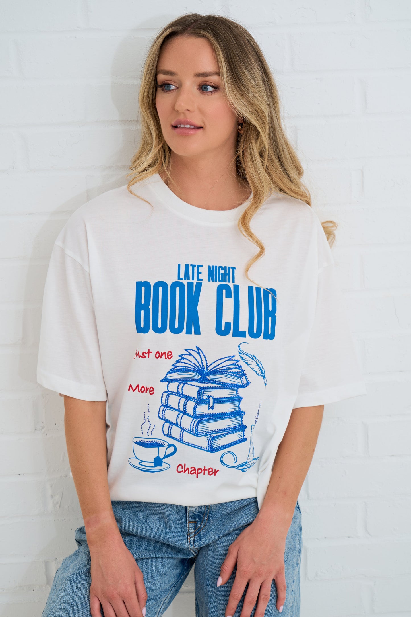 Woman wearing a white t-shirt with 'Late Night Book Club' design against a white brick wall.