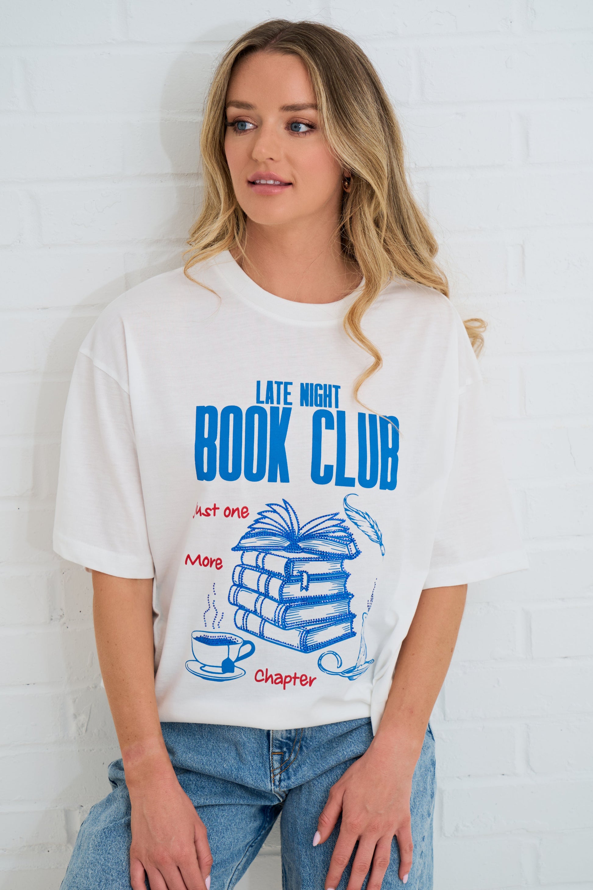 Woman wearing a white t-shirt with 'Late Night Book Club' design against a white brick wall.