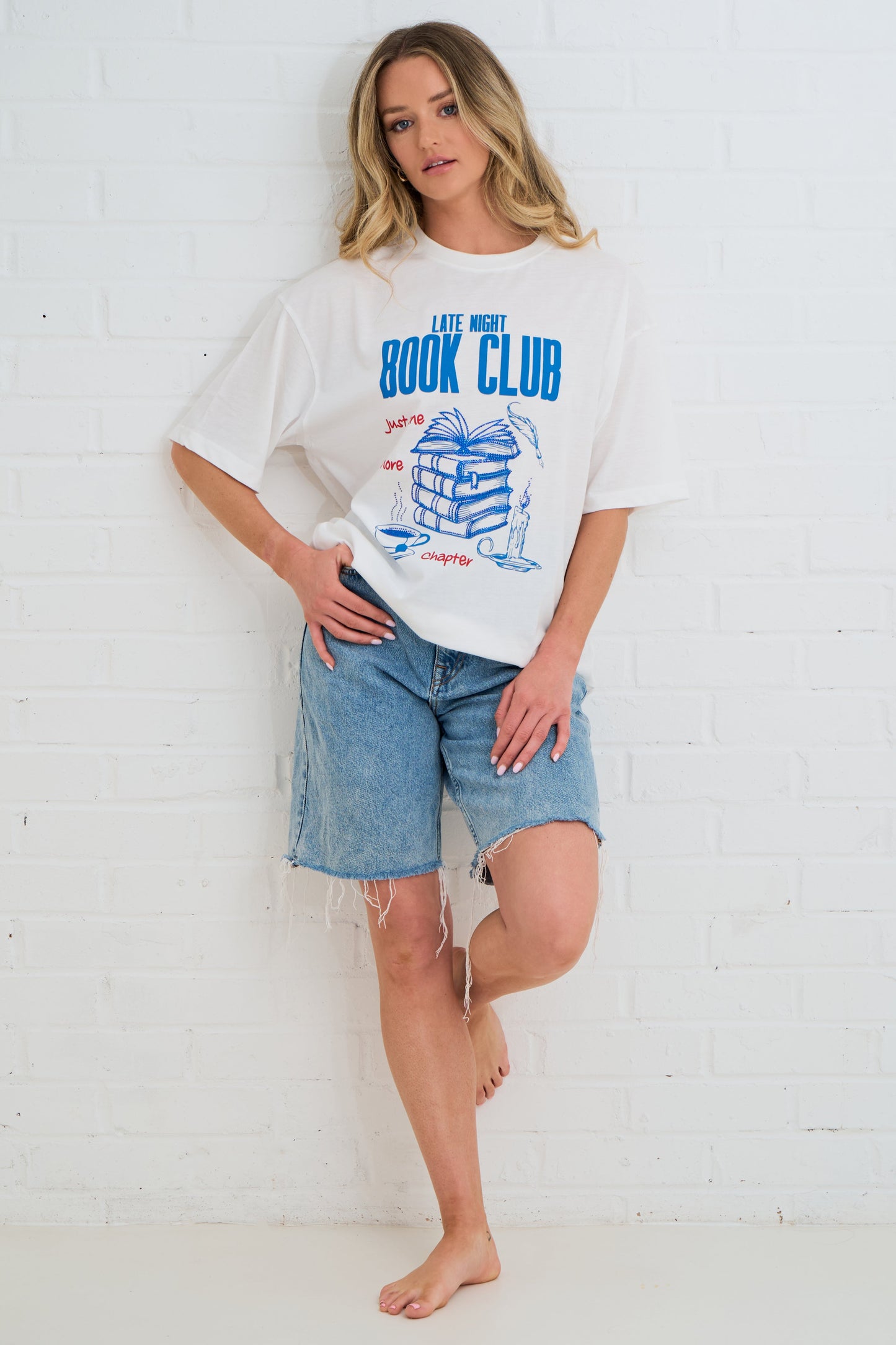 Woman wearing a white t-shirt with 'Late Night Book Club' design and denim shorts against a white brick wall.