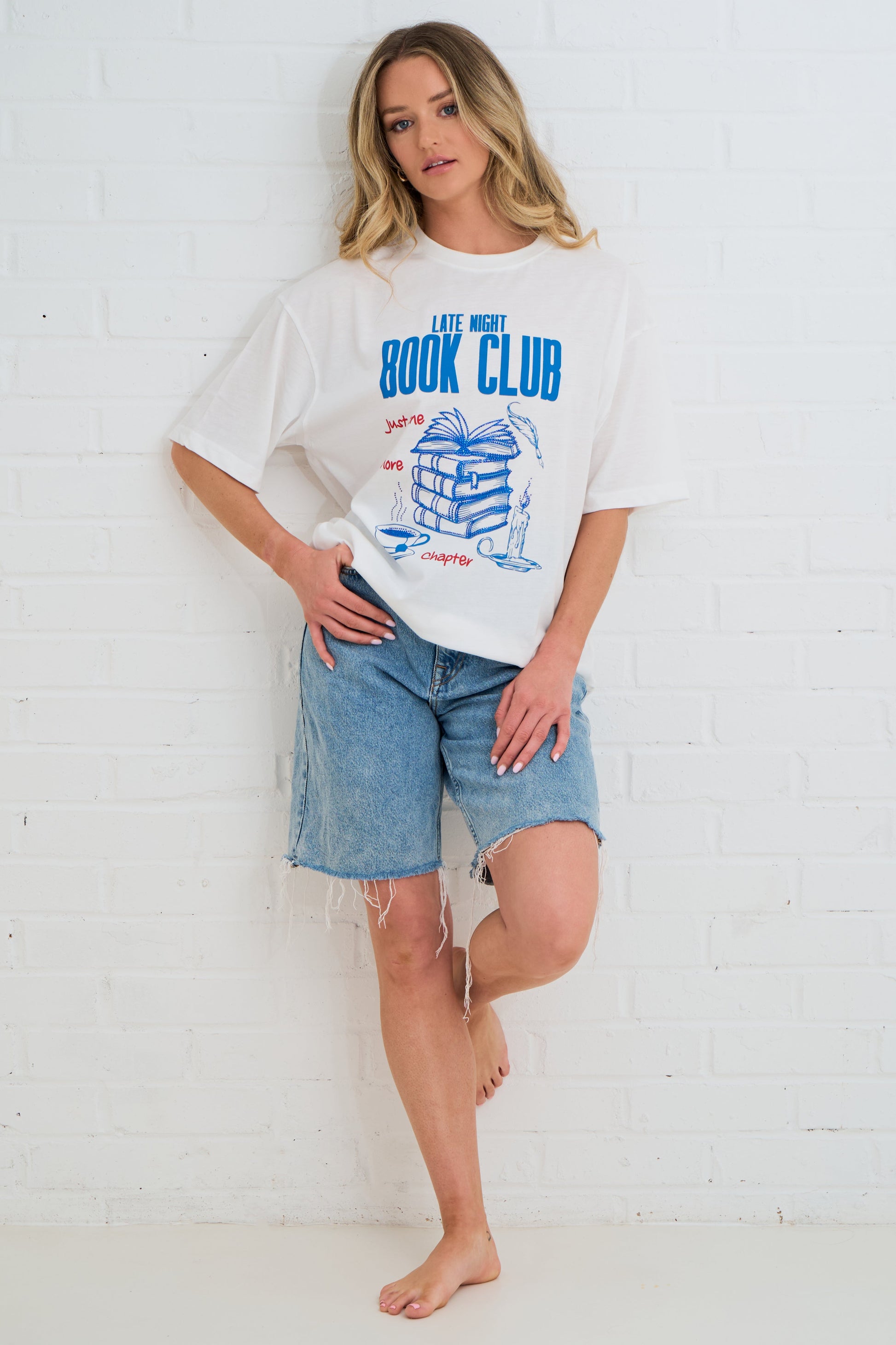 Woman wearing a white t-shirt with 'Late Night Book Club' design and denim shorts against a white brick wall.