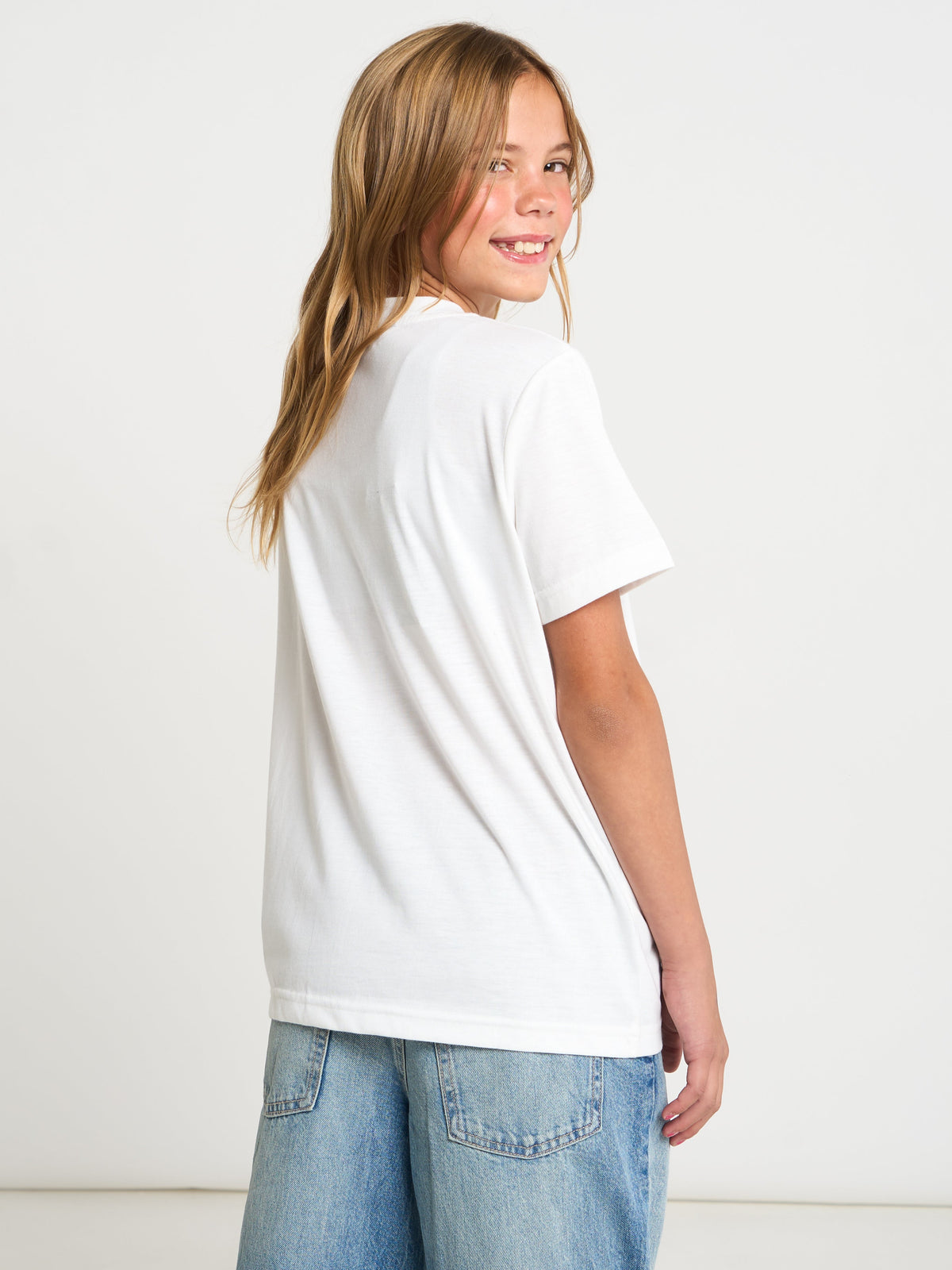 A young girl with long blonde hair is shown from the back wearing a plain white short-sleeve t-shirt and light blue denim jeans against a clean white studio background.